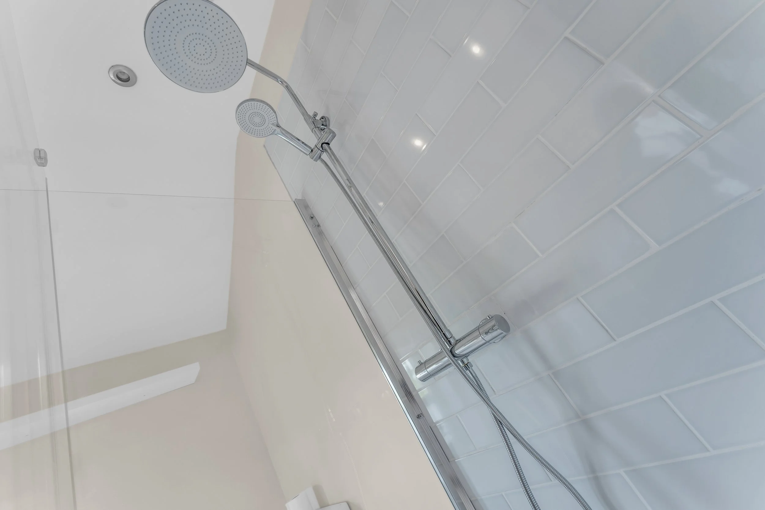 A modern shower area featuring two chrome shower heads on a tiling wall. The wall has gray tiles, and the shower heads are attached to a horizontal bar with a flexible hose.