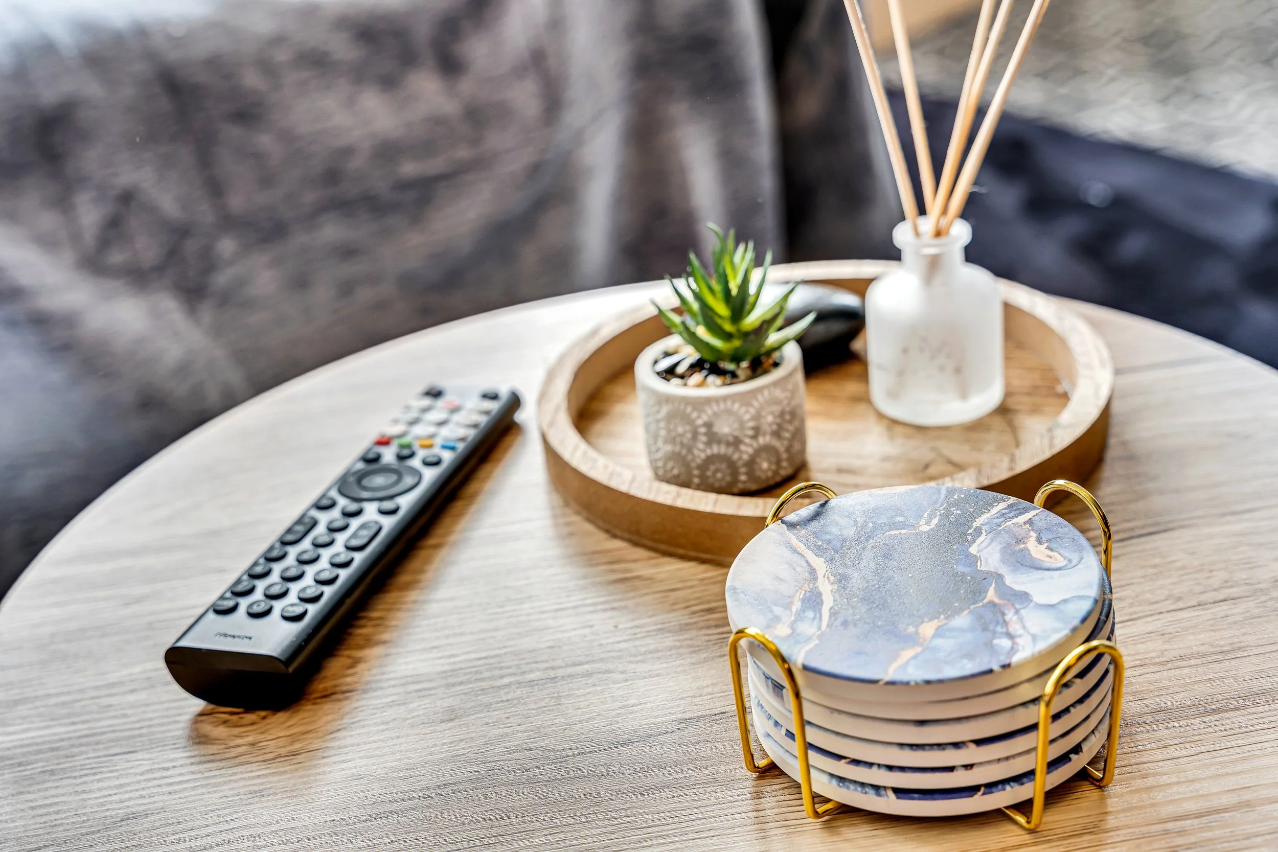 A wooden table with a remote control, a small potted succulent, a white vase with reed diffusers, and a set of coasters with a marble pattern, all arranged on a round wooden tray.
