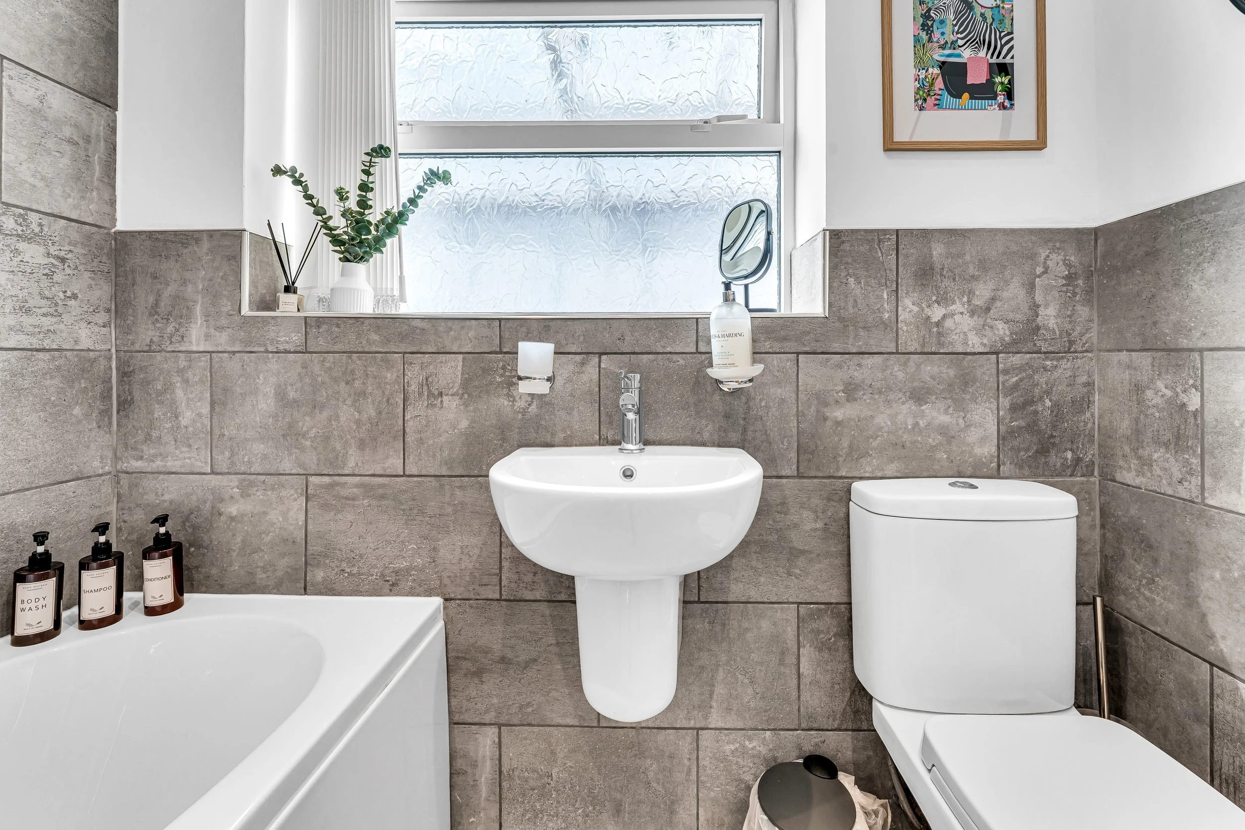 Modern bathroom with a window, a wall-mounted sink, a toilet, a bathtub with toiletries, a potted plant, soap dispenser, other bathroom decor, and a framed picture on the wall.