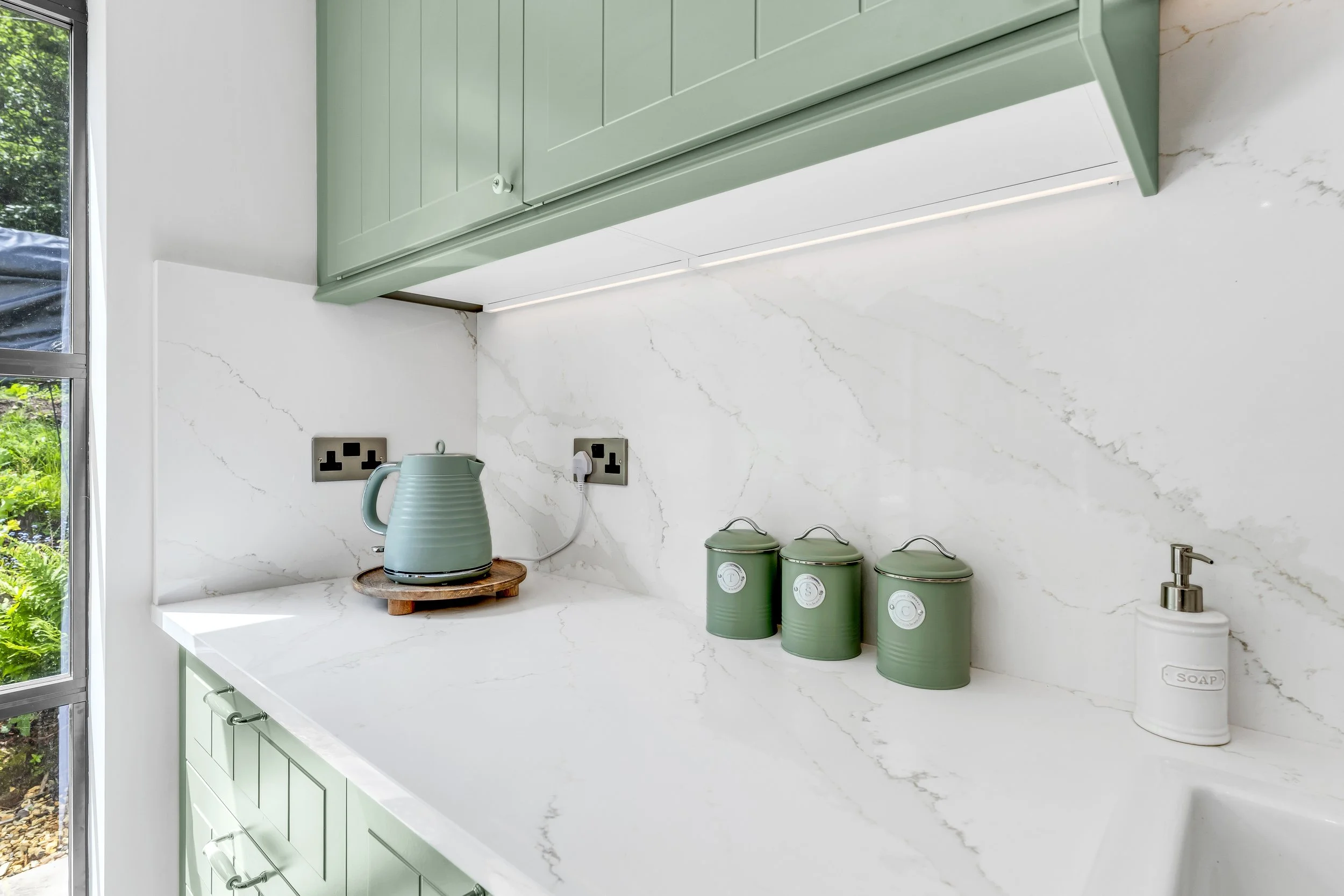 A section of a modern kitchen with white marble countertops and a marble backsplash, green kitchen cabinets, a window with greenery outside, and kitchen accessories including a teal kettle on a wooden stand, three matching green containers with white
