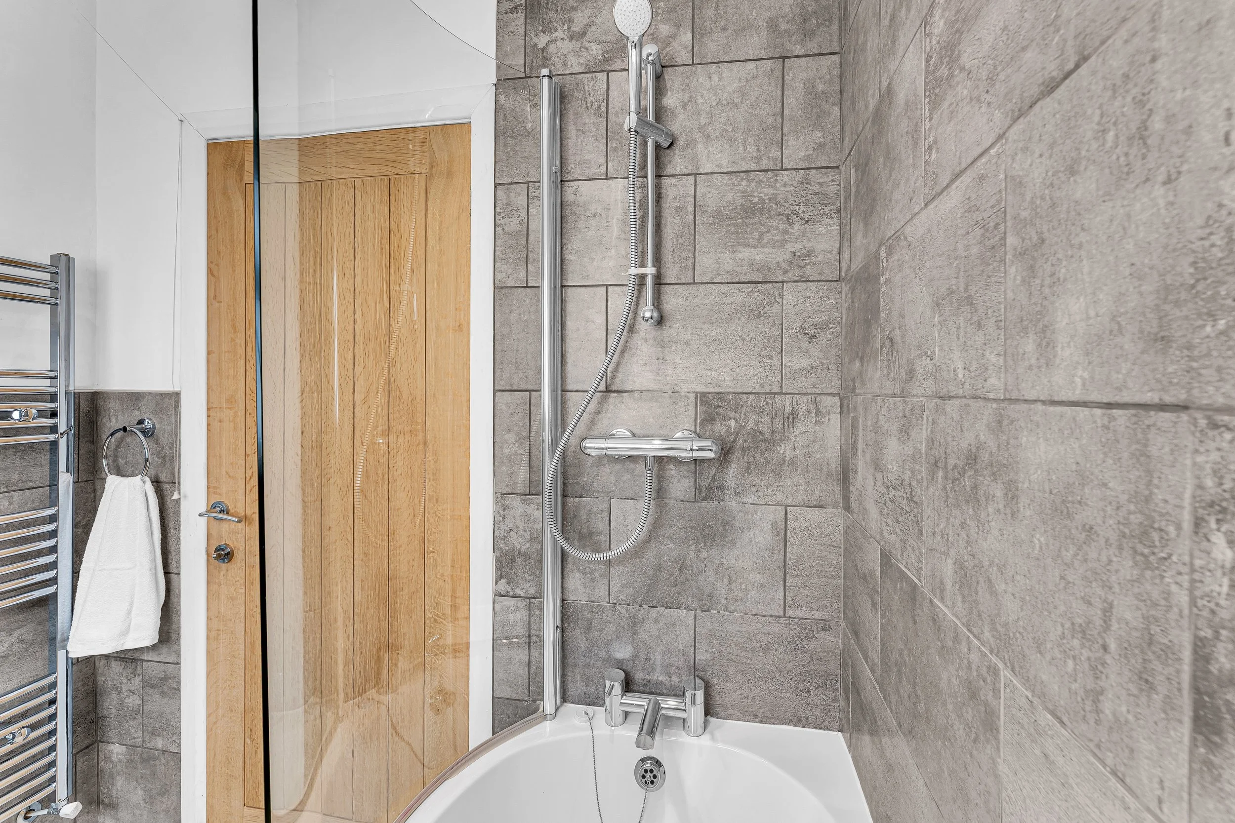 Modern bathroom with gray tiled wall, bathtub with chrome fixtures, and a showerhead mounted on a sliding rail. A wooden door and towel rack with a white towel are also visible.