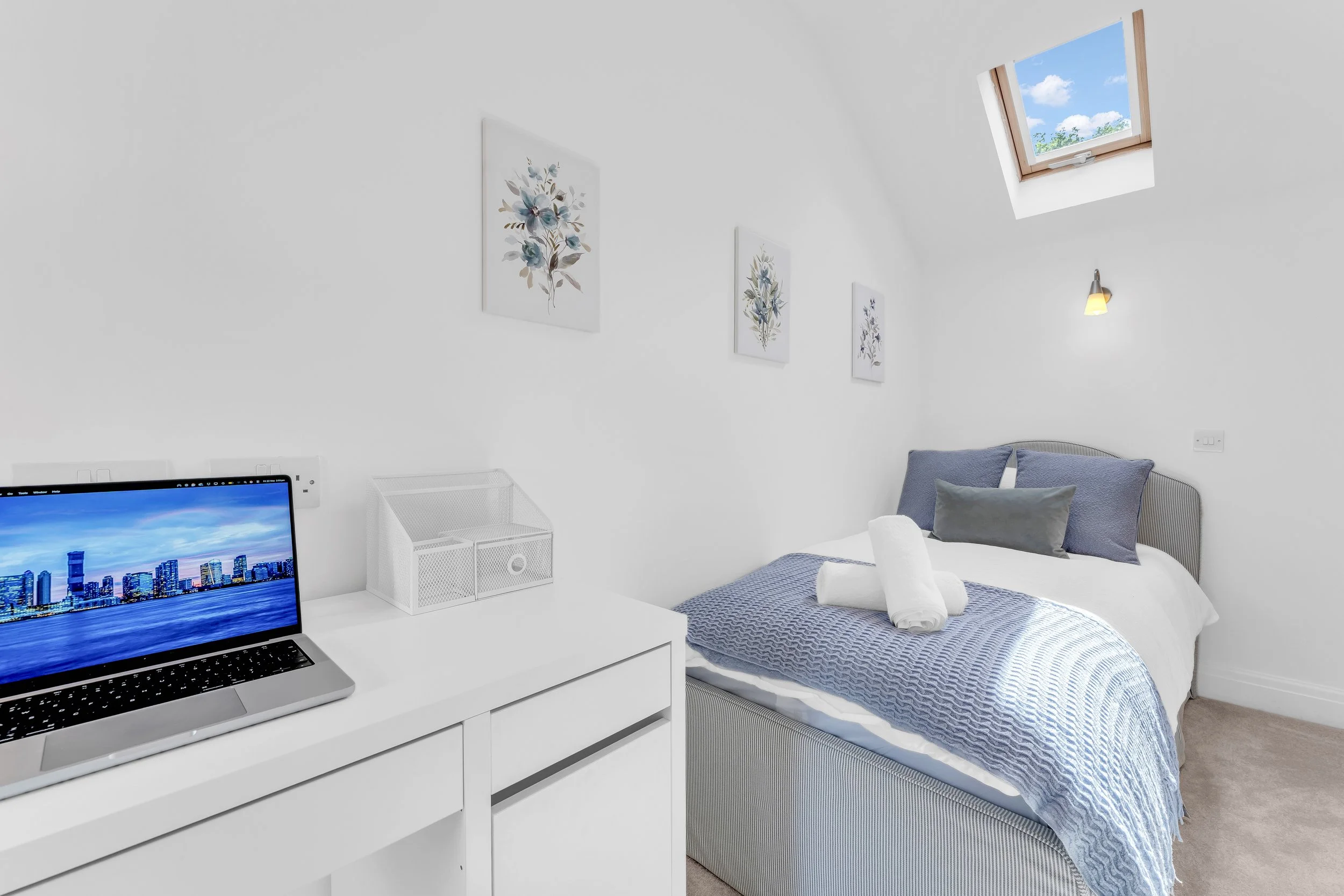 A white bedroom with a skylight showing a blue sky with clouds. The bed has a grey and white striped headboard, a grey blanket and a grey pillow. A towel is on the bed. On the wall above, there are three floral art prints. A small wall-mounted light 