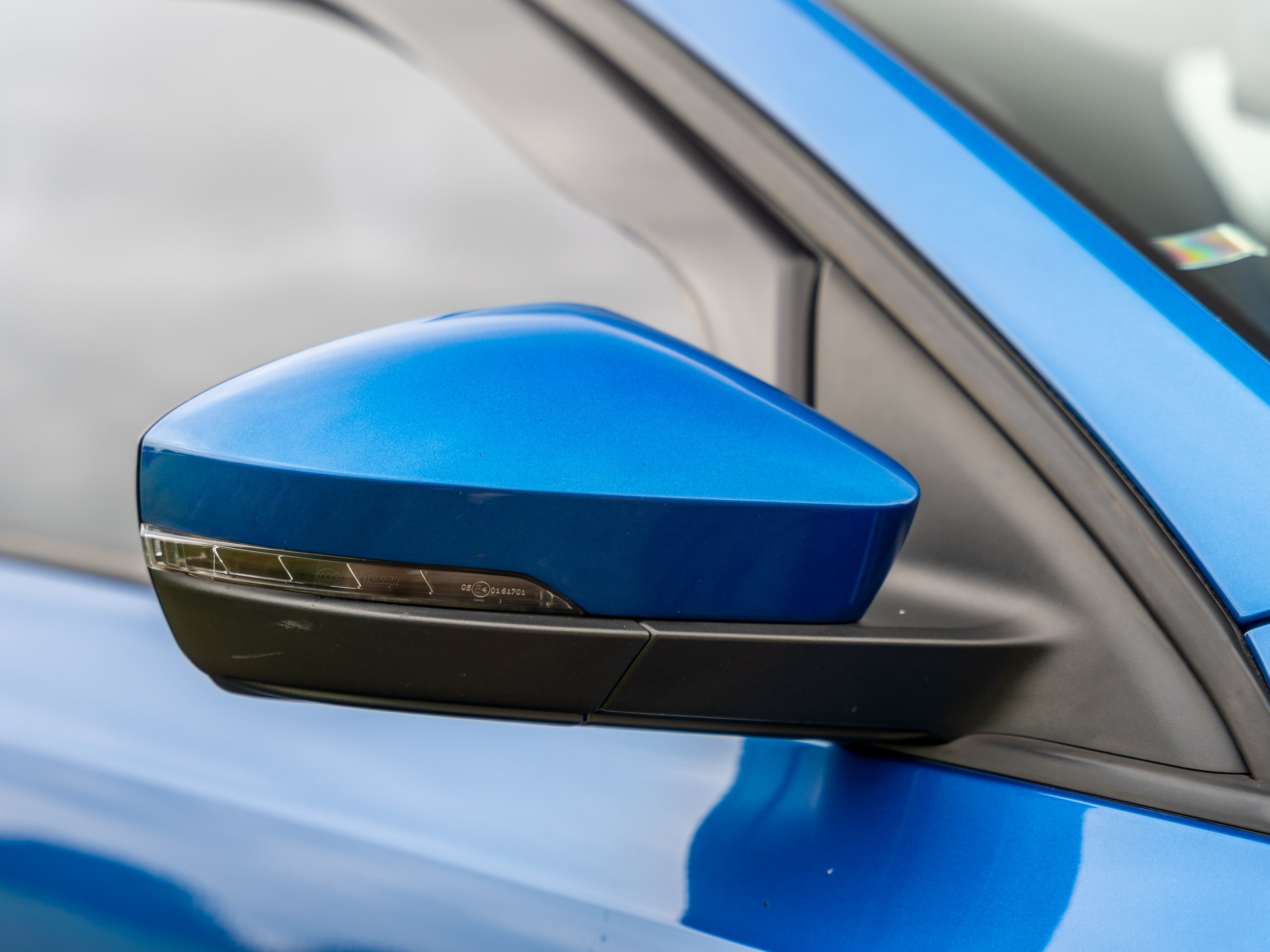 Close-up of a blue car side mirror with a black base and integrated turn signal, part of the car's blue body and windshield are also visible.
