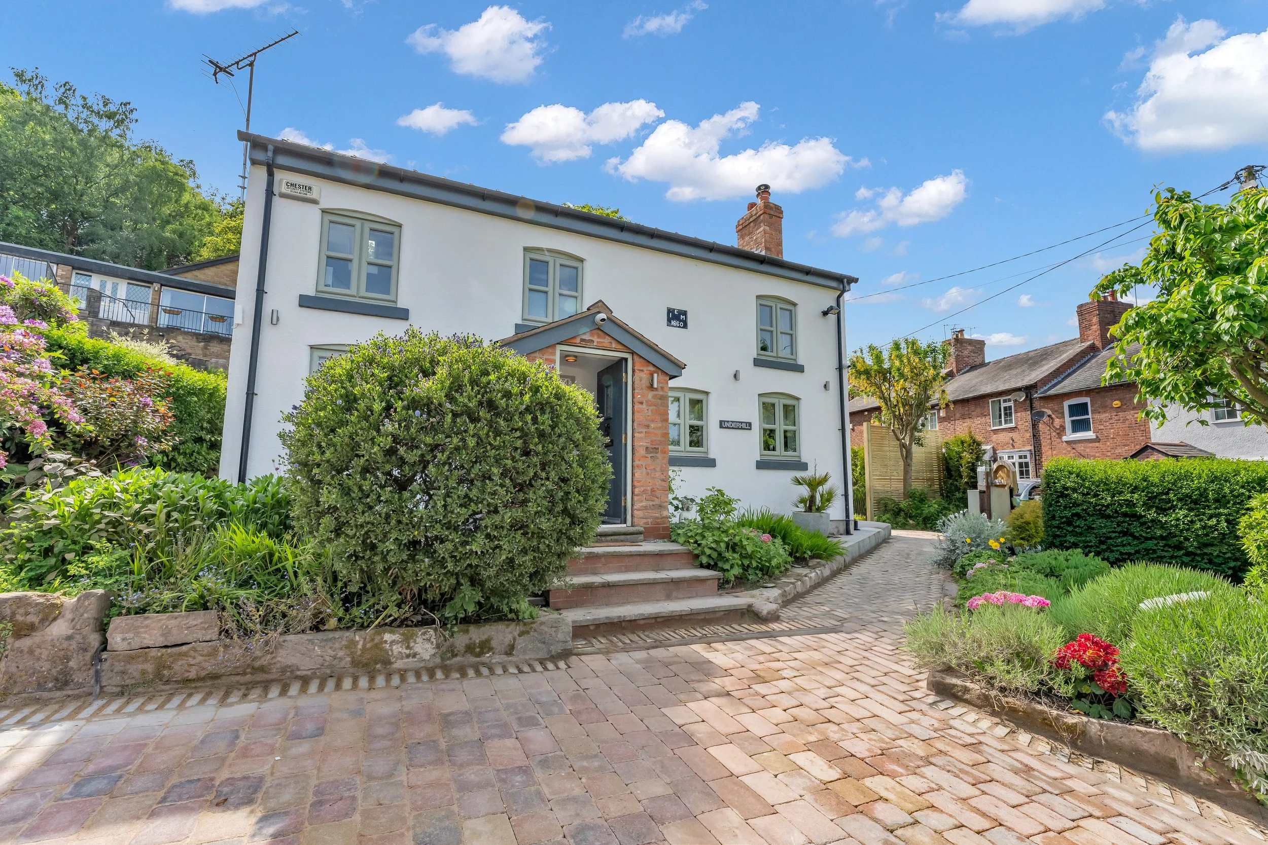 A two-story white house with green windows and brick accents, surrounded by a lush garden with colorful flowers and bushes, a cobblestone pathway, and a clear blue sky with scattered clouds.