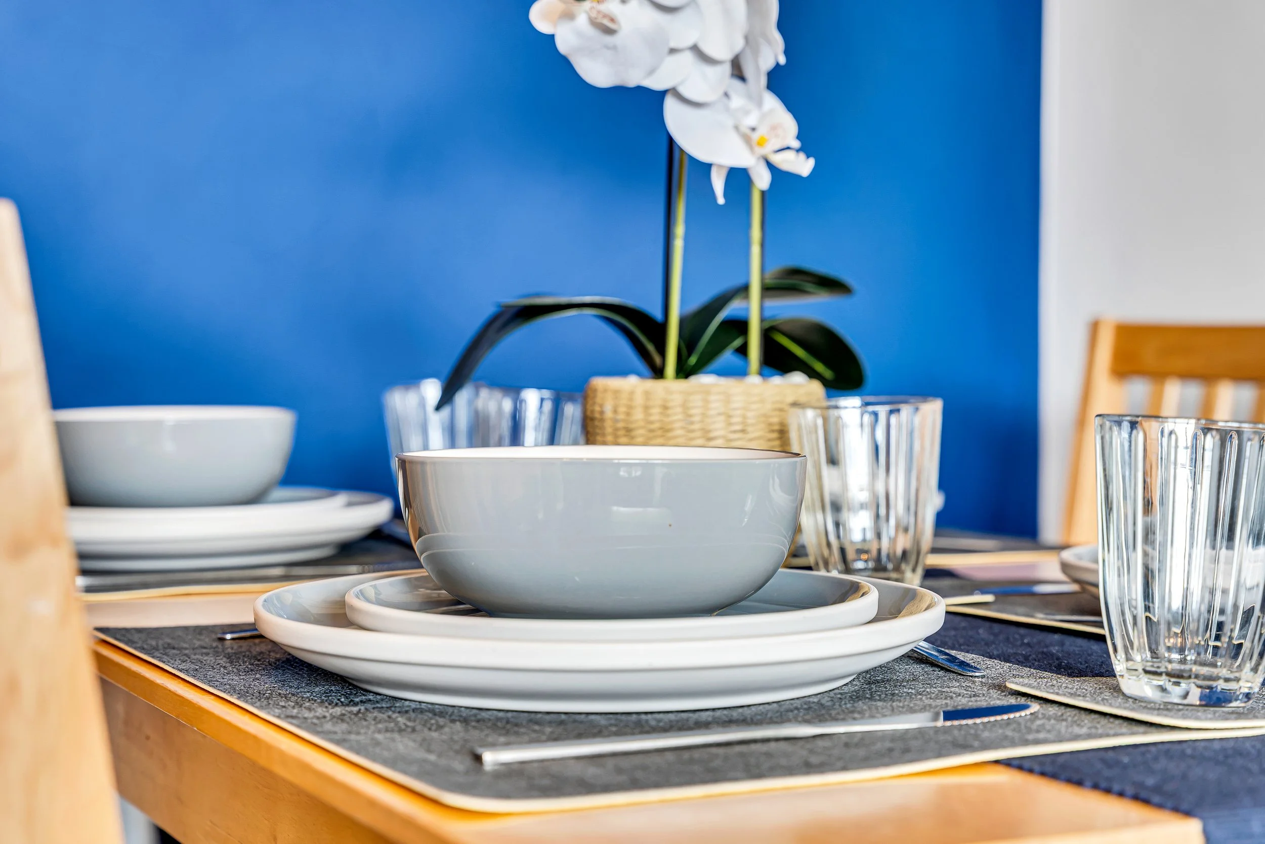 A close-up of a dining table with white plates, bowls, clear glass tumblers, and silverware, with a white orchid plant in the background against a blue wall.