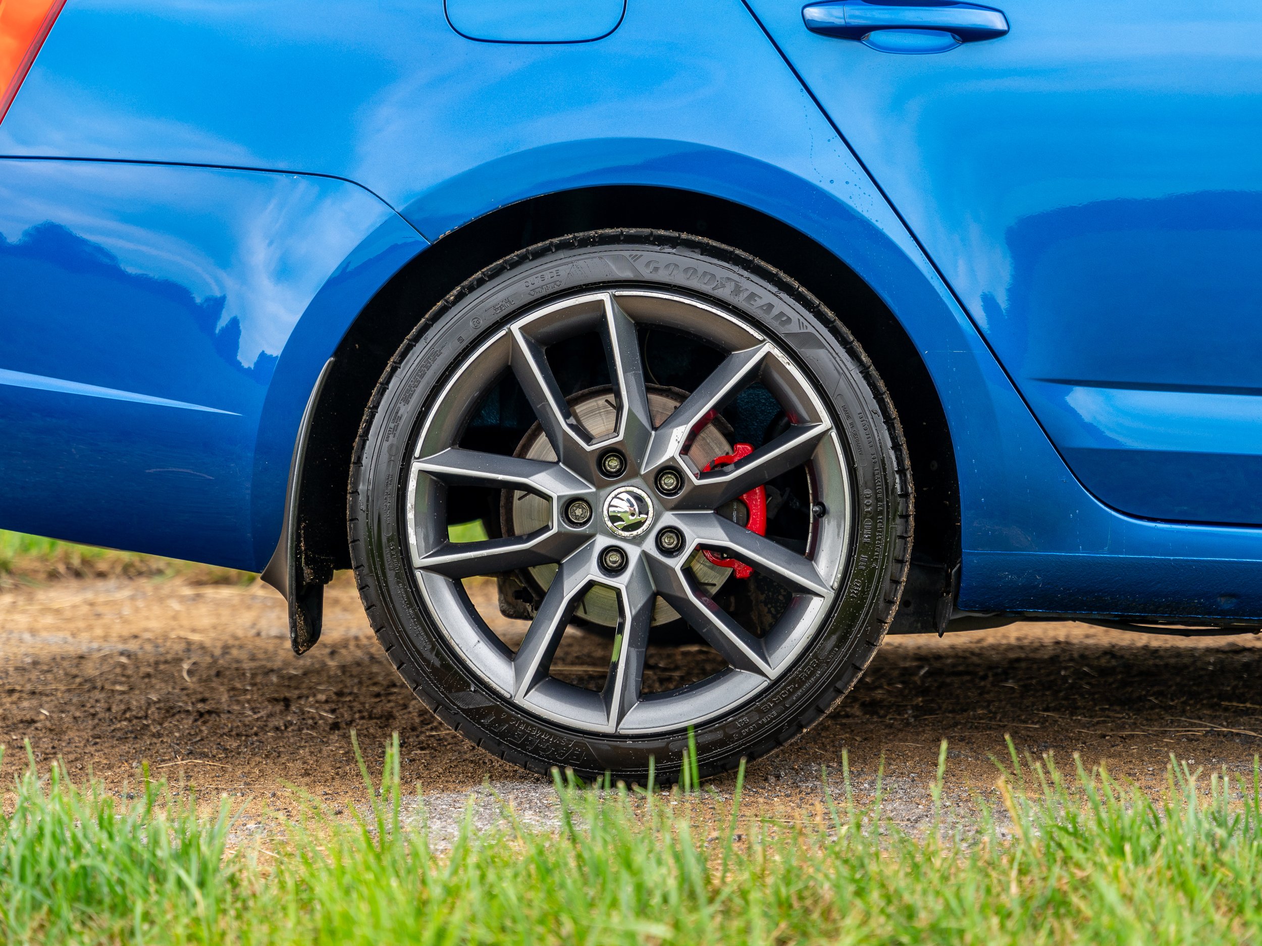 Close-up of a blue car's front right wheel with a multi-spoke alloy rim, a red brake caliper, and a Goodyear tire, parked on a grass and dirt surface.