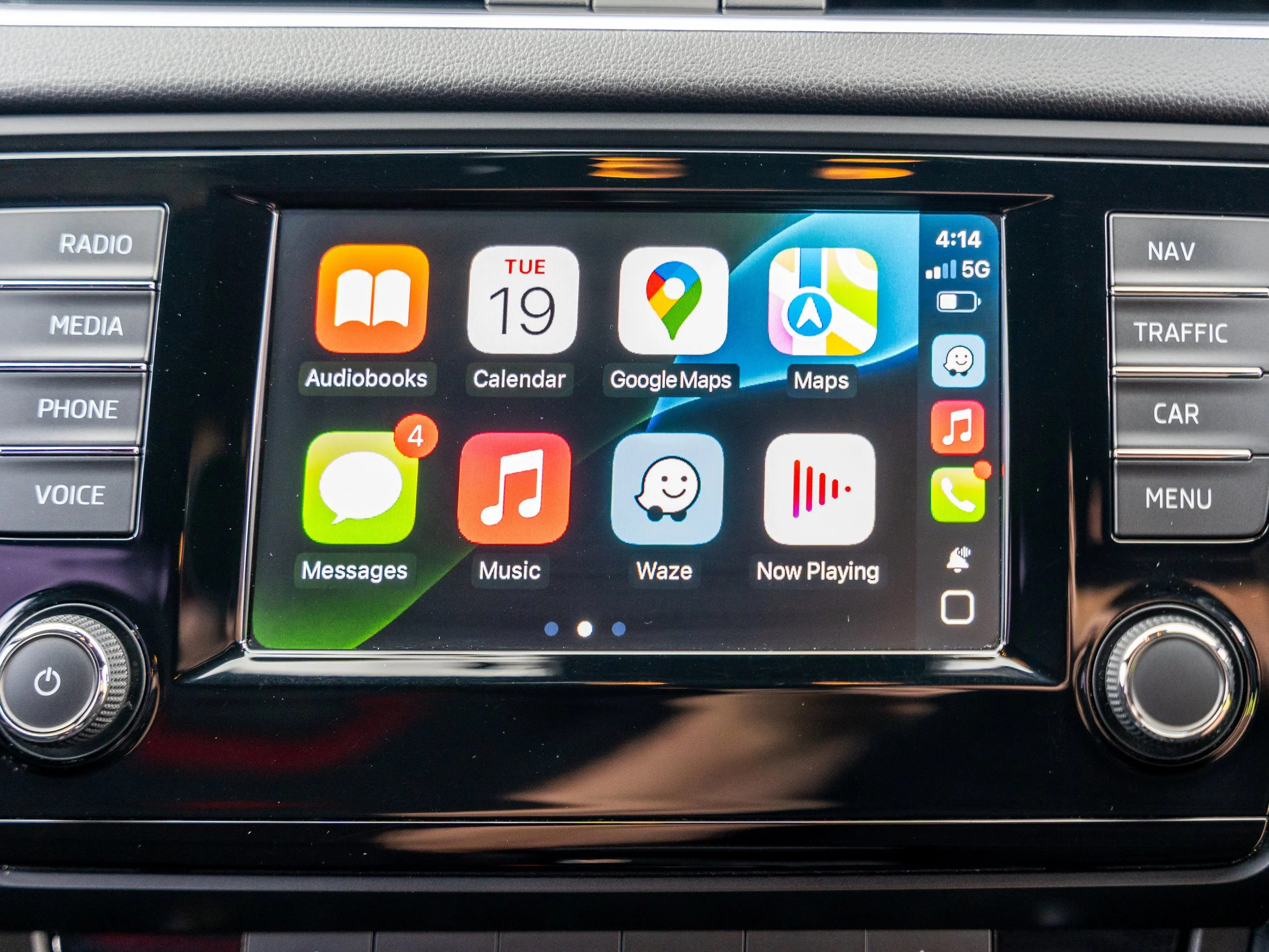 Car infotainment touchscreen displaying apps such as Audiobooks, Calendar, Google Maps, Maps, Messages, Music, Waze, and Now Playing, with physical buttons on the sides labeled RADIO, MEDIA, PHONE, VOICE, NAV, TRAFFIC, CAR, and MENU.