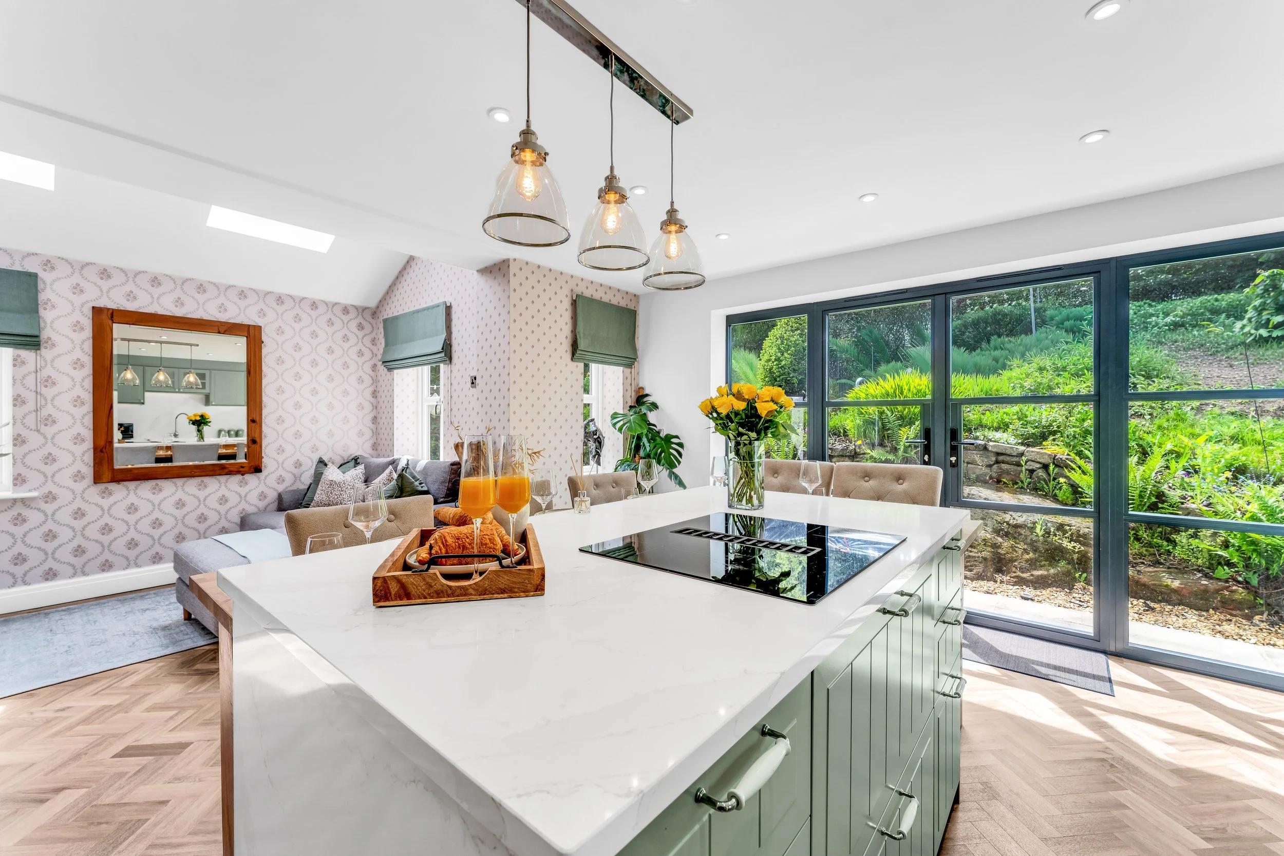 Modern kitchen with white quartz island, green cabinets, large windows revealing greenery outside, and pendant lights hanging from ceiling.