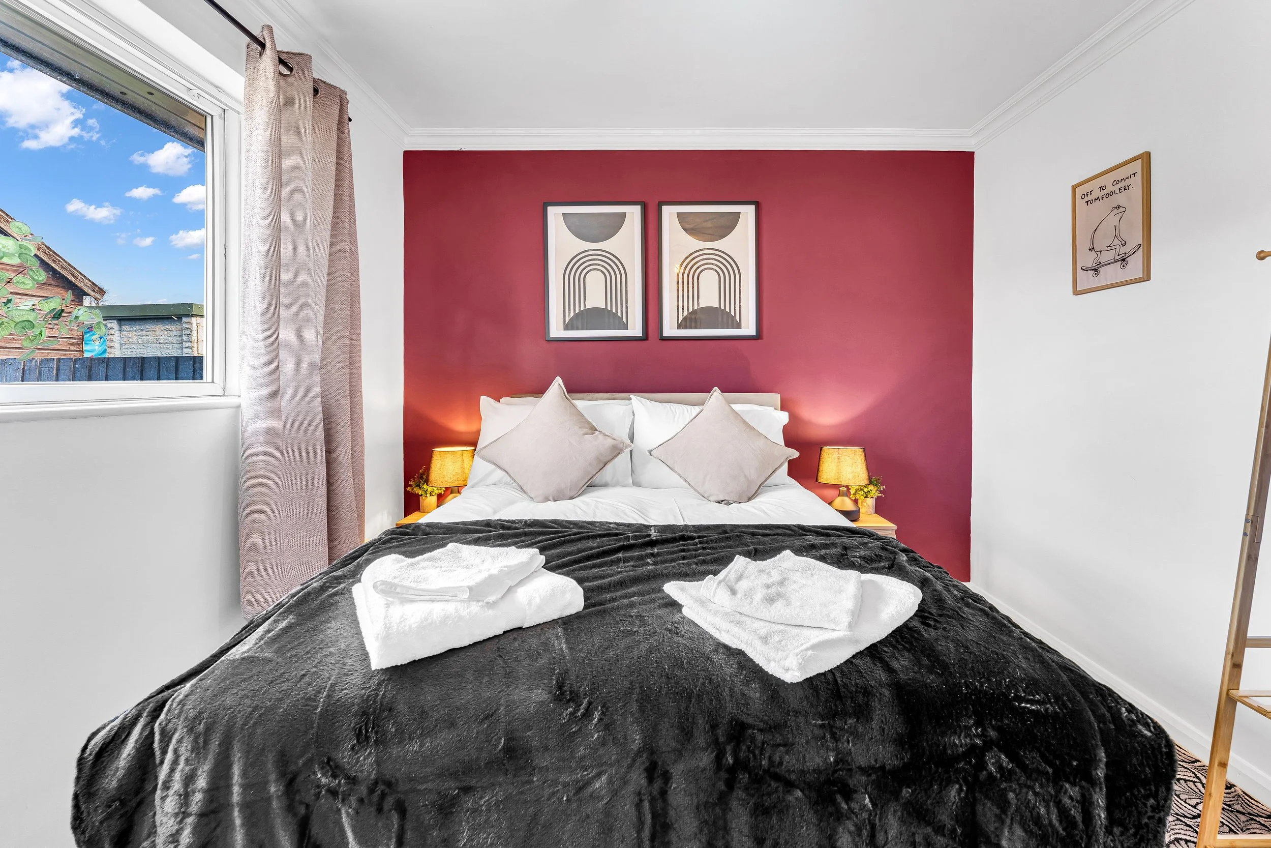 A bedroom with a bed featuring white pillows and a dark gray blanket, two bedside lamps, and towels on the bed. The wall behind the bed is painted red with two framed abstract art pieces, and there is a window with light-colored curtains on the left 