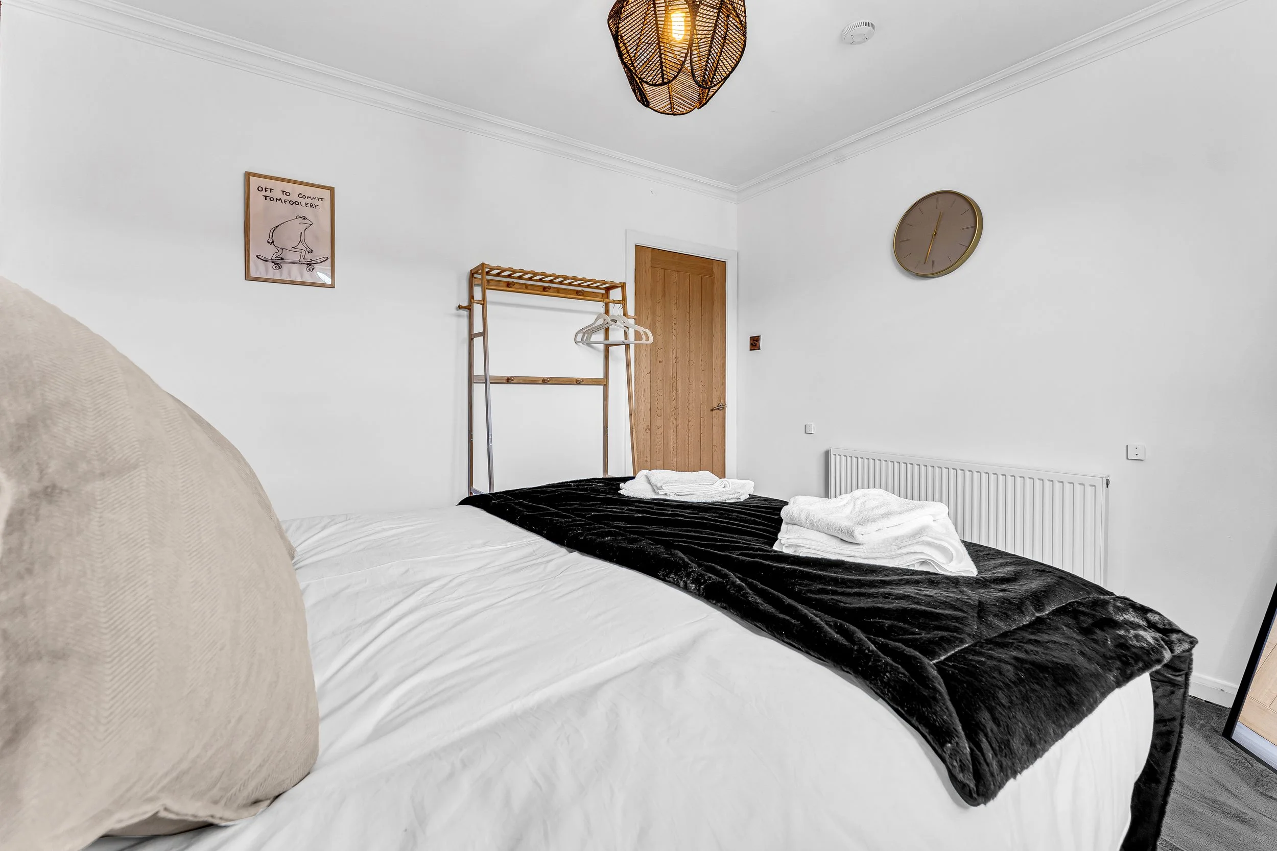 Bedroom with a bed covered in white sheets and a black blanket, two folded white towels on the bed, beige cushion on the bed corner, wooden door, round wall clock, framed wall art, wooden clothing rack with hangers, ceiling light fixture, white radia