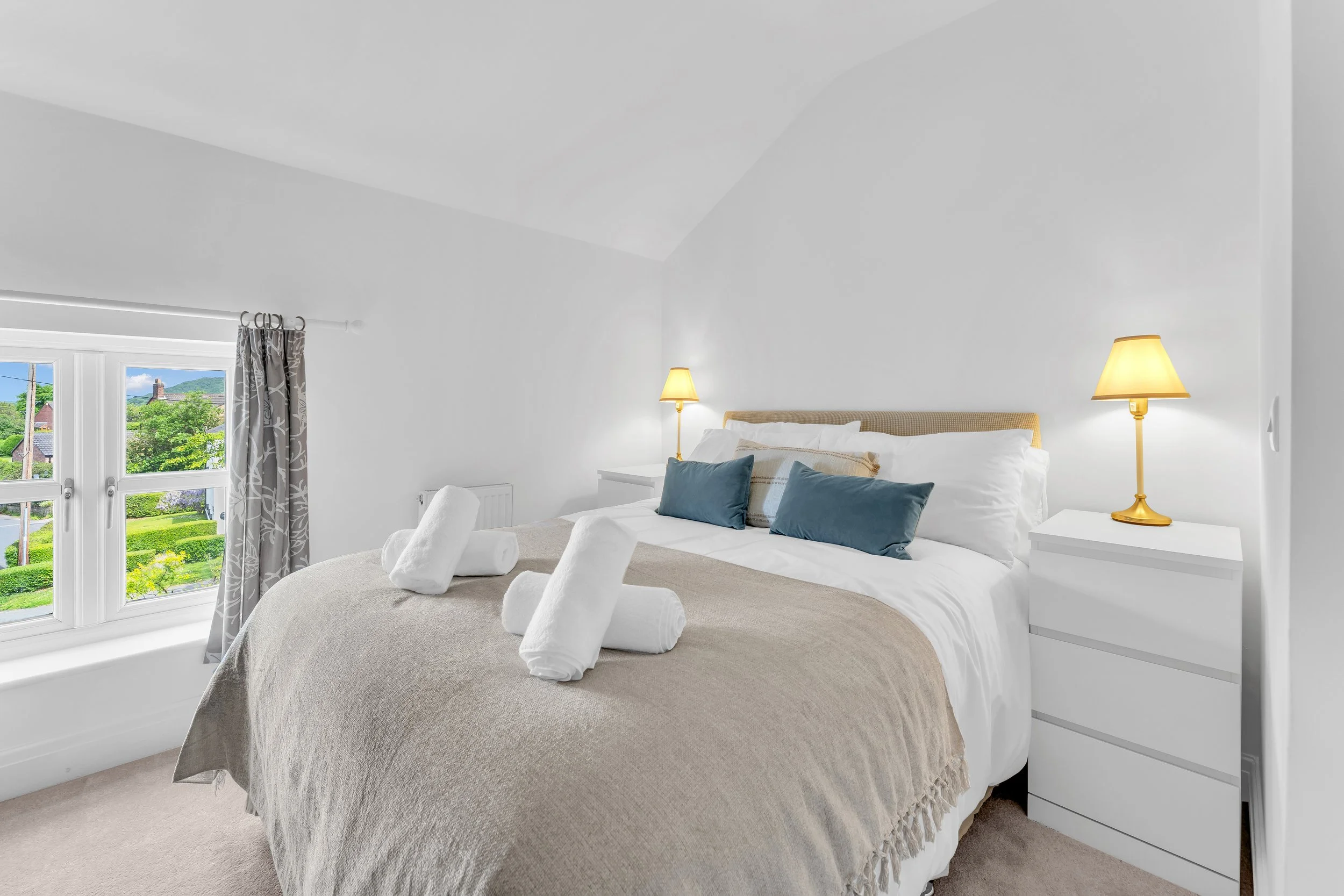 A cozy bedroom with a large bed covered in a beige blanket and adorned with white and blue pillows. Four white towels are rolled and placed at the foot of the bed. The room has white walls, a white dresser, and two yellow bedside lamps. There is a wi