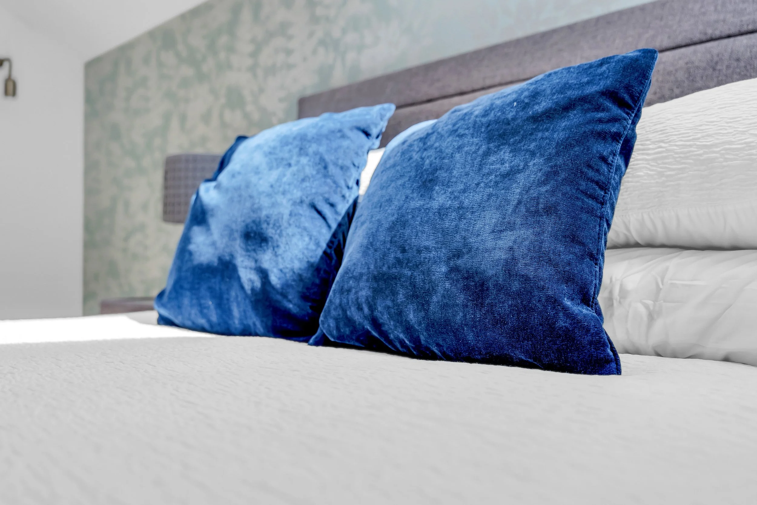 Close-up of a bed with two blue velvet pillows and a white textured blanket, headboard with a gray fabric, and a patterned wall in the background.