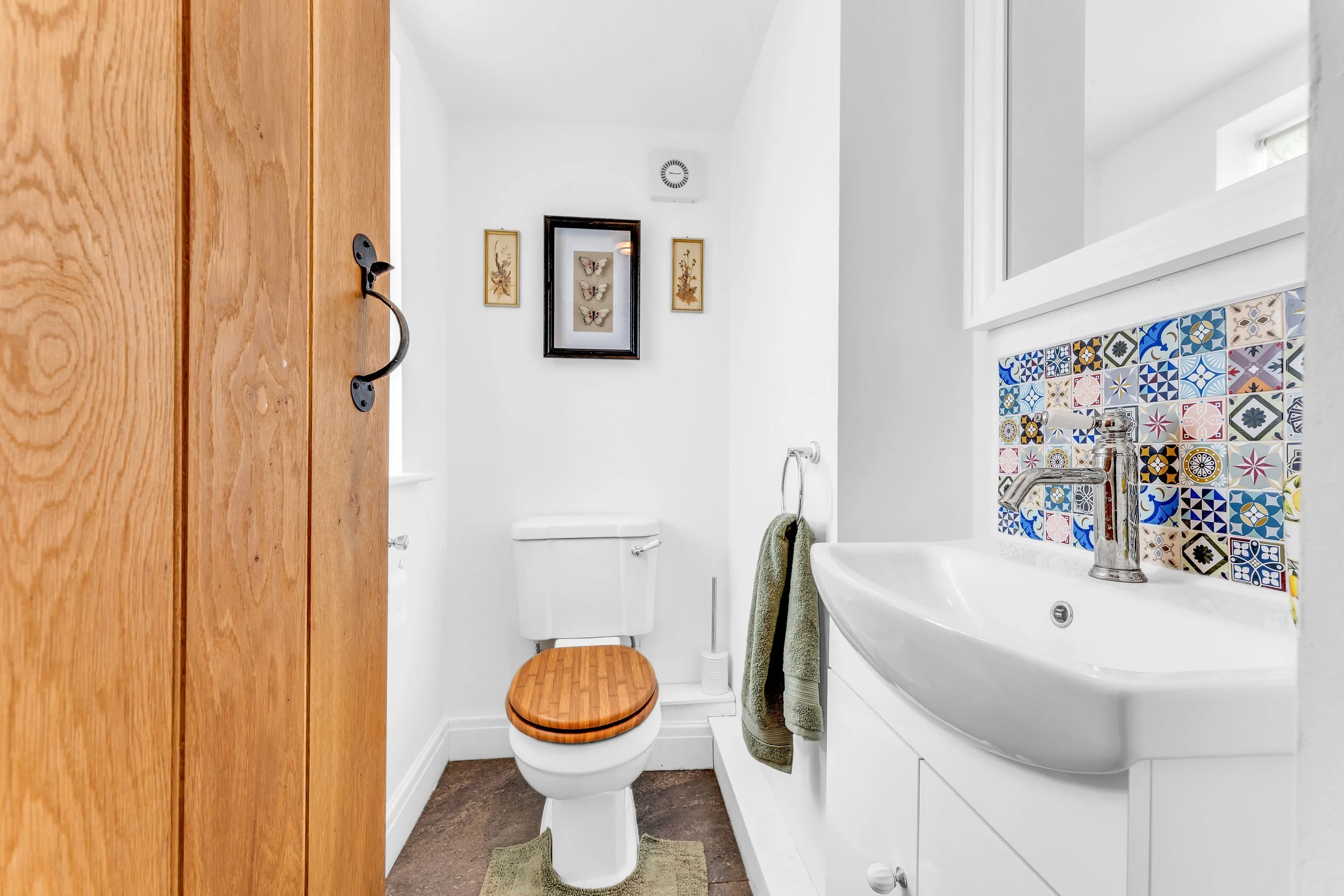 Small bathroom with white walls, wooden door, and decorative tiles behind the sink. Features a toilet, small sink with chrome faucet, green towels, and framed butterfly and floral artwork on the wall.
