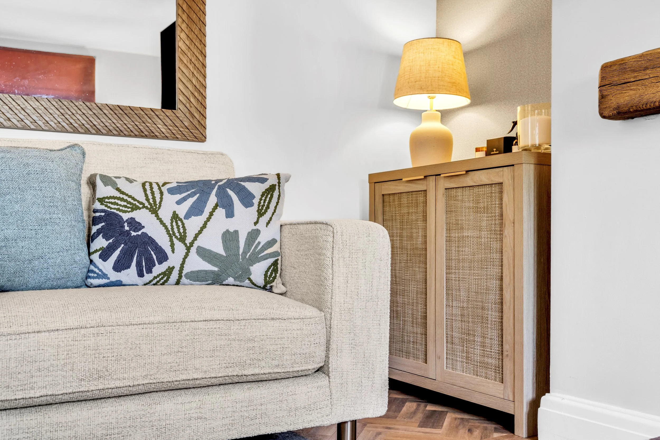 Close-up of a beige upholstered sofa with decorative pillows, a wooden cabinet with rattan doors, a table lamp with a beige shade, and a framed mirror on the wall in a cozy living room.