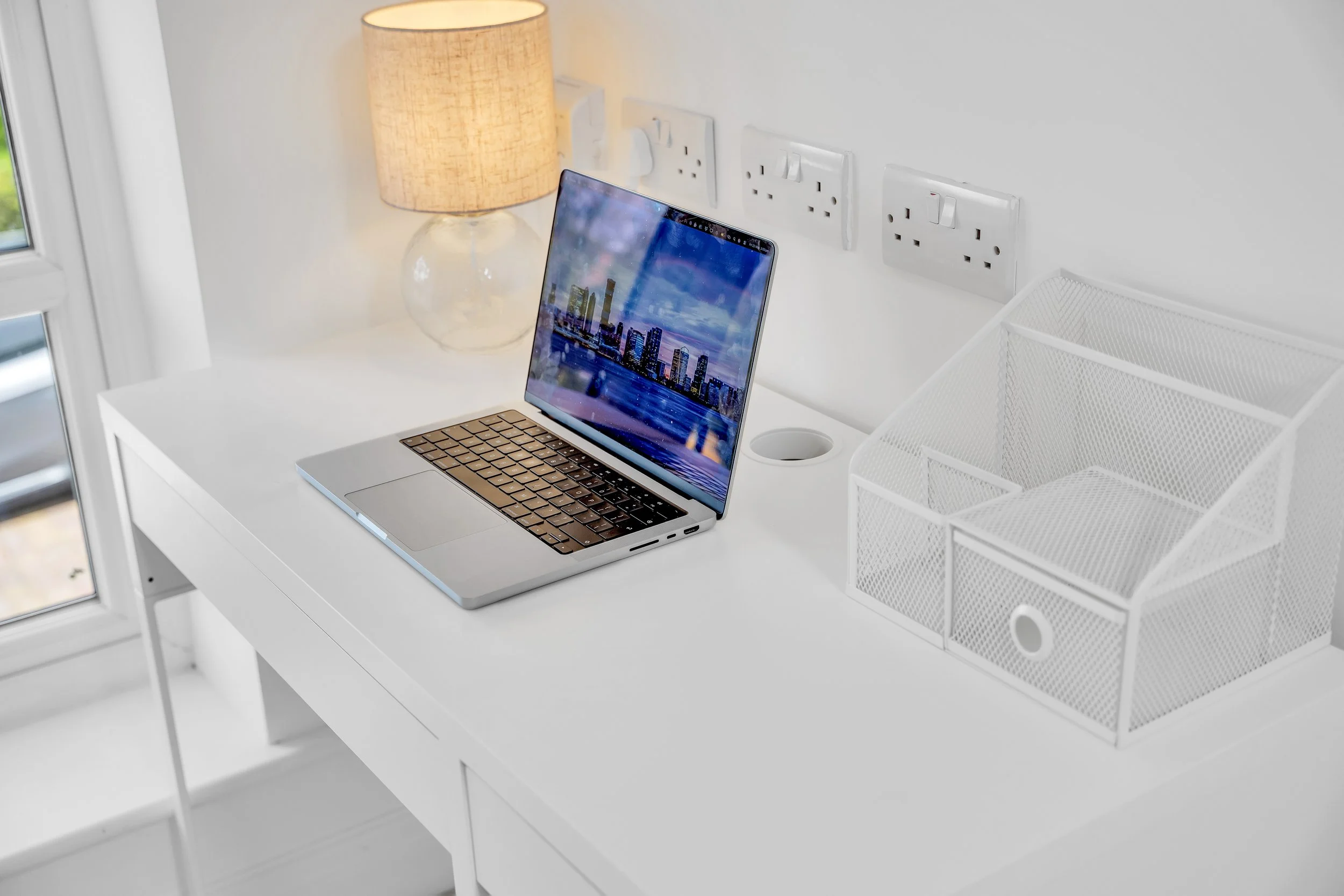 White desk with a silver laptop displaying a city skyline, a beige table lamp, and a white wire organizer, near a wall with power outlets. A window is partially visible on the left.