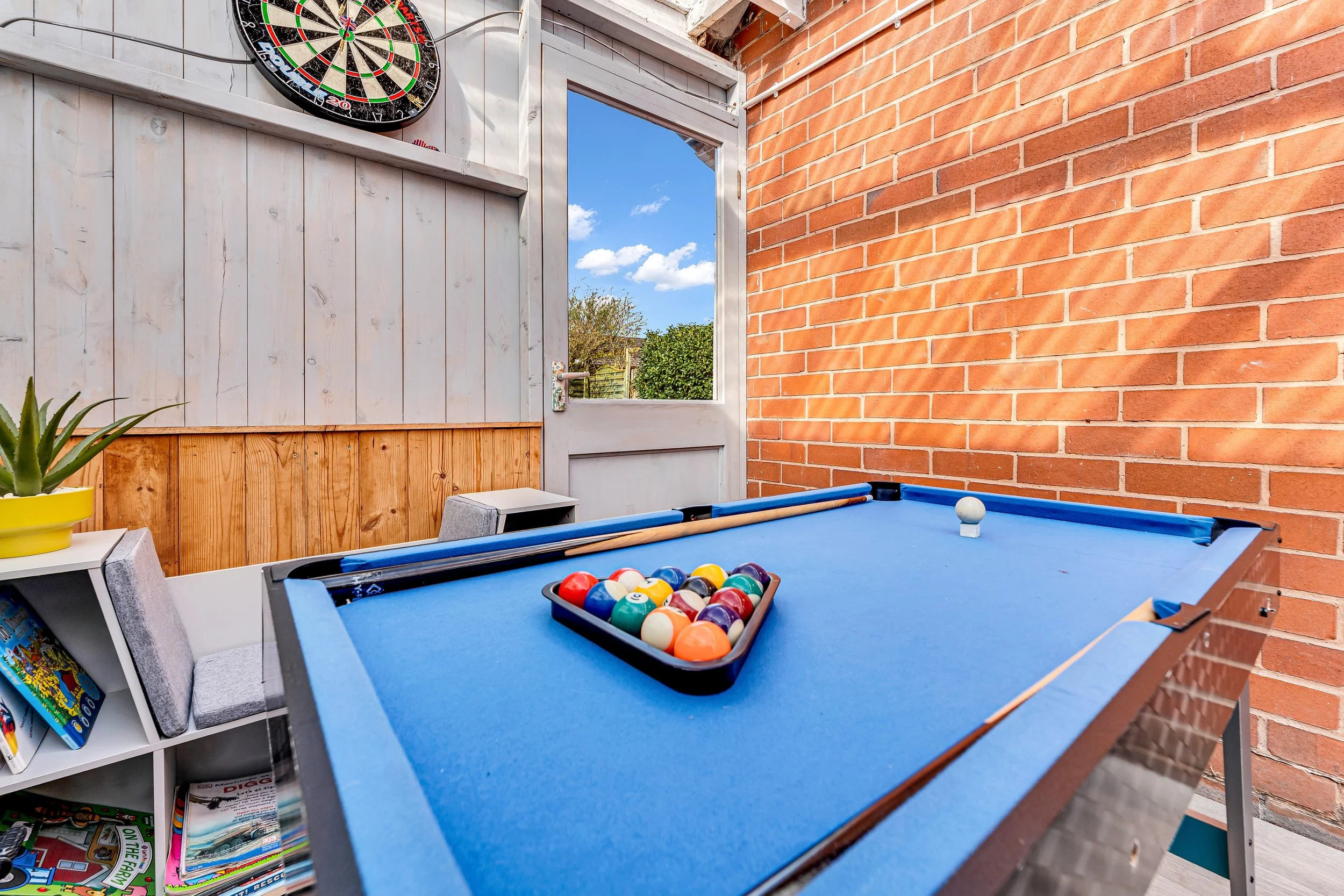 An outdoor game room with a blue pool table, a tray of billiard balls, a cue stick, and an air-hockey table mount on a brick wall. Outside view through an open door shows blue sky with clouds and green trees.