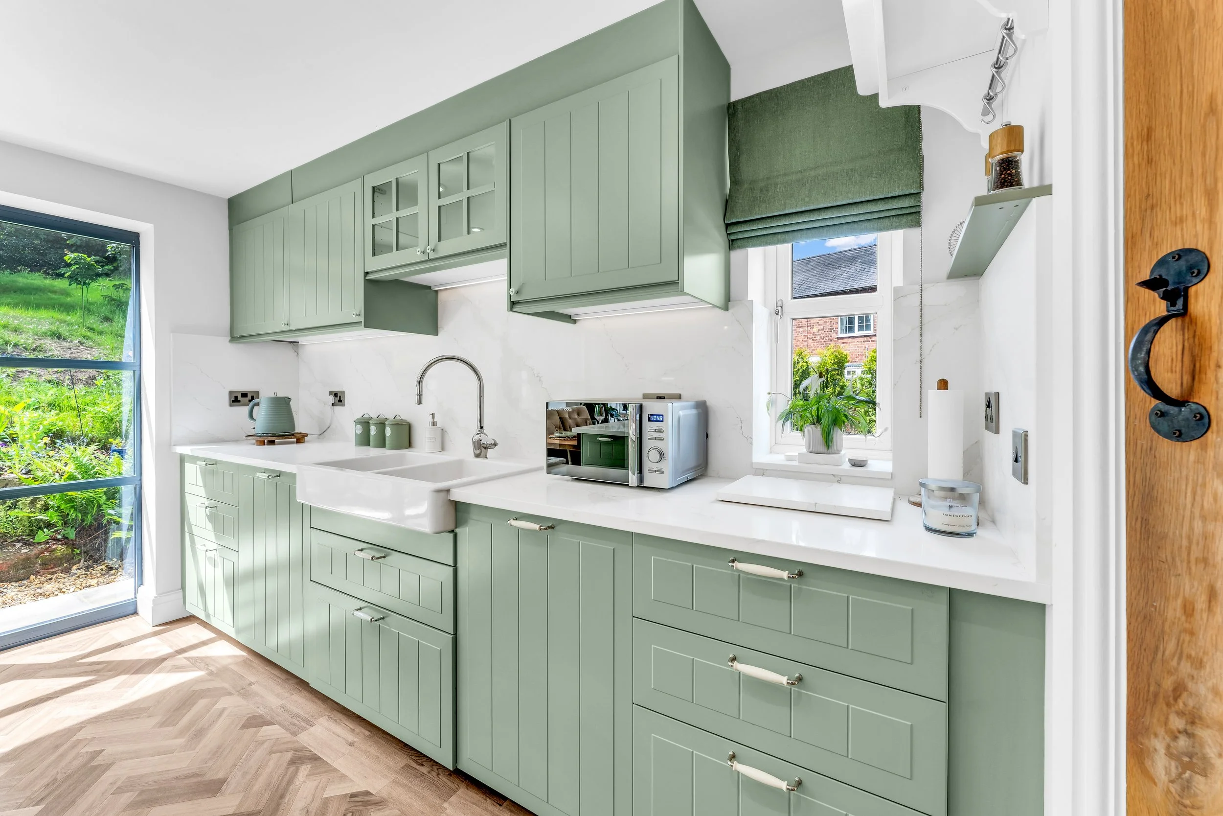 A bright kitchen with green cabinets, white countertops, a white sink, a microwave, and a window with green Roman shades revealing a garden outside.