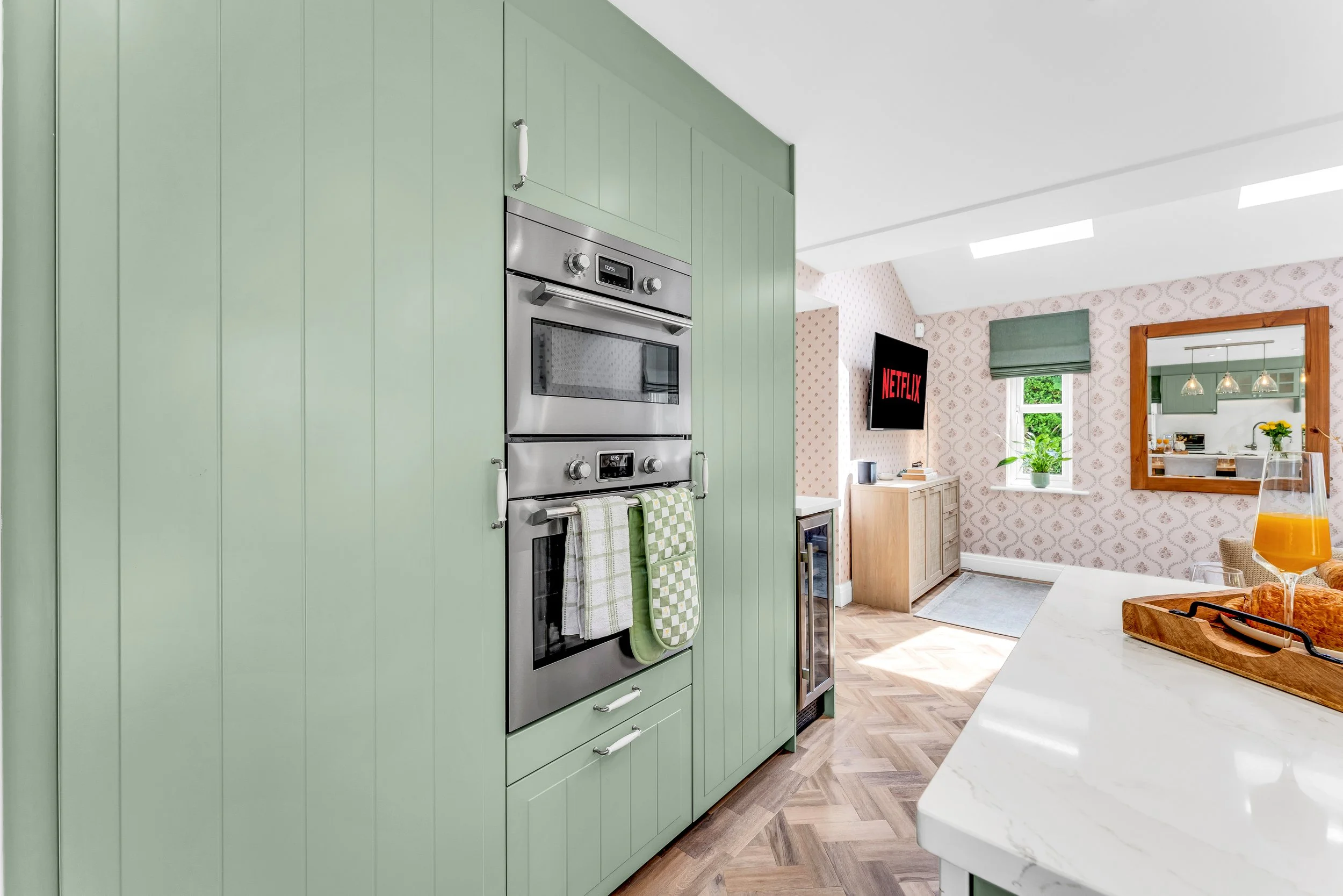 A modern kitchen with mint green cabinets, stainless steel oven, and a view into a dining area with floral wallpaper, a window with a green Roman shade, and a mirror reflecting pendant lights and a dining table.