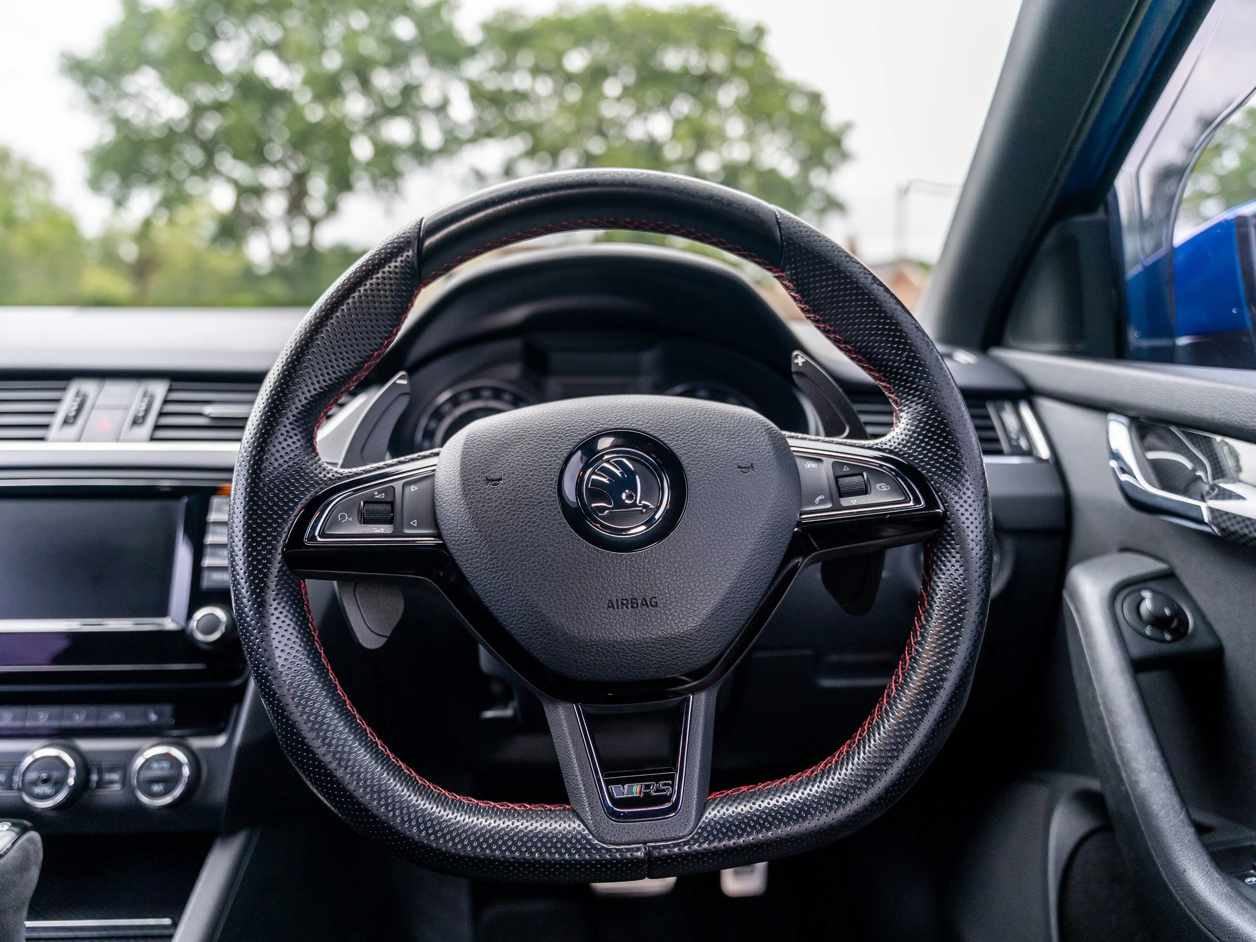 Close-up of a steering wheel in a car's interior, showing the dashboard and controls, with trees outside through the windshield.