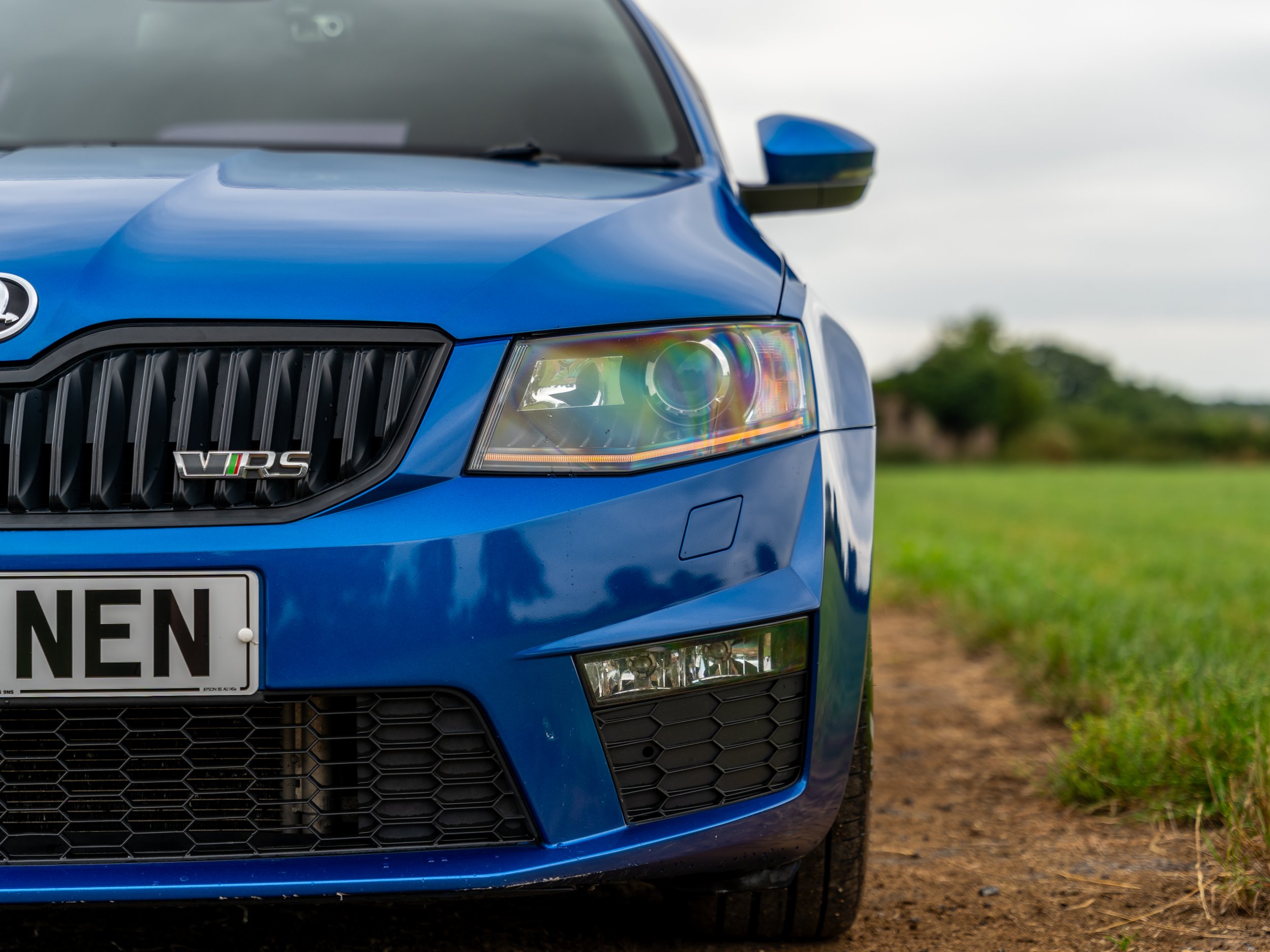 Close-up of the front of a blue Skoda VRS car parked on a dirt road, with fields and trees in the background.