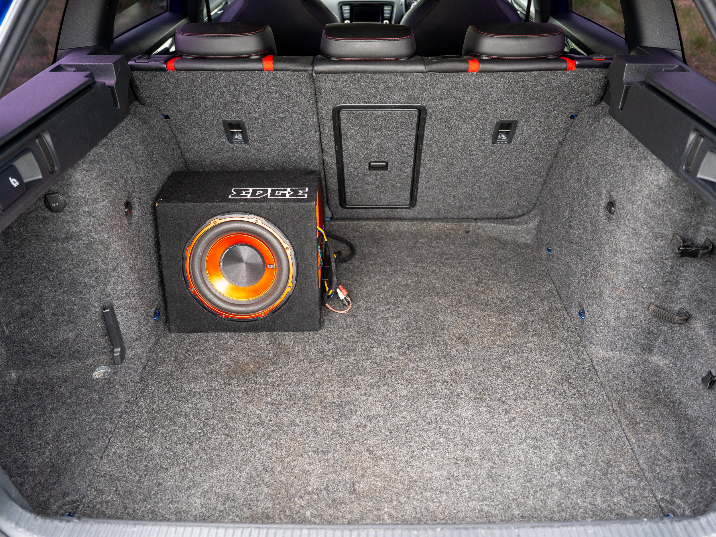 Empty car trunk with a subwoofer speaker installed on the left side, gray carpet lining, rear seats with headrests visible, and car interior details.