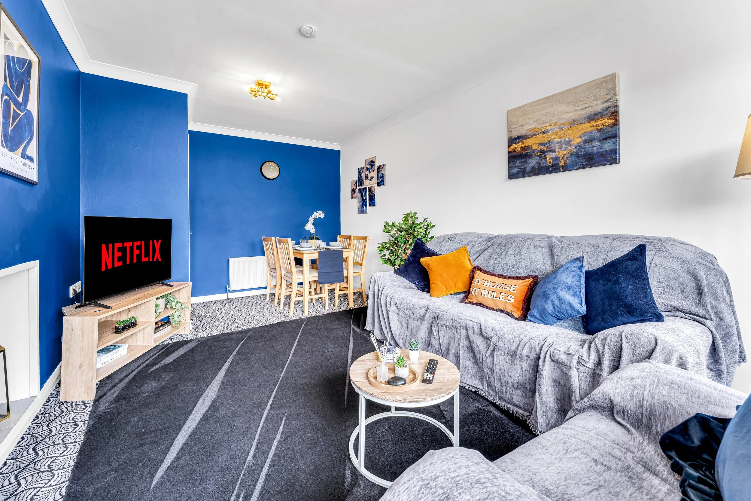 Living room with gray sofas, blue and orange pillows, a coffee table with small plants and remote, a TV displaying Netflix, a dining area with a table and wooden chairs, blue accent wall with artwork, and decorative items.