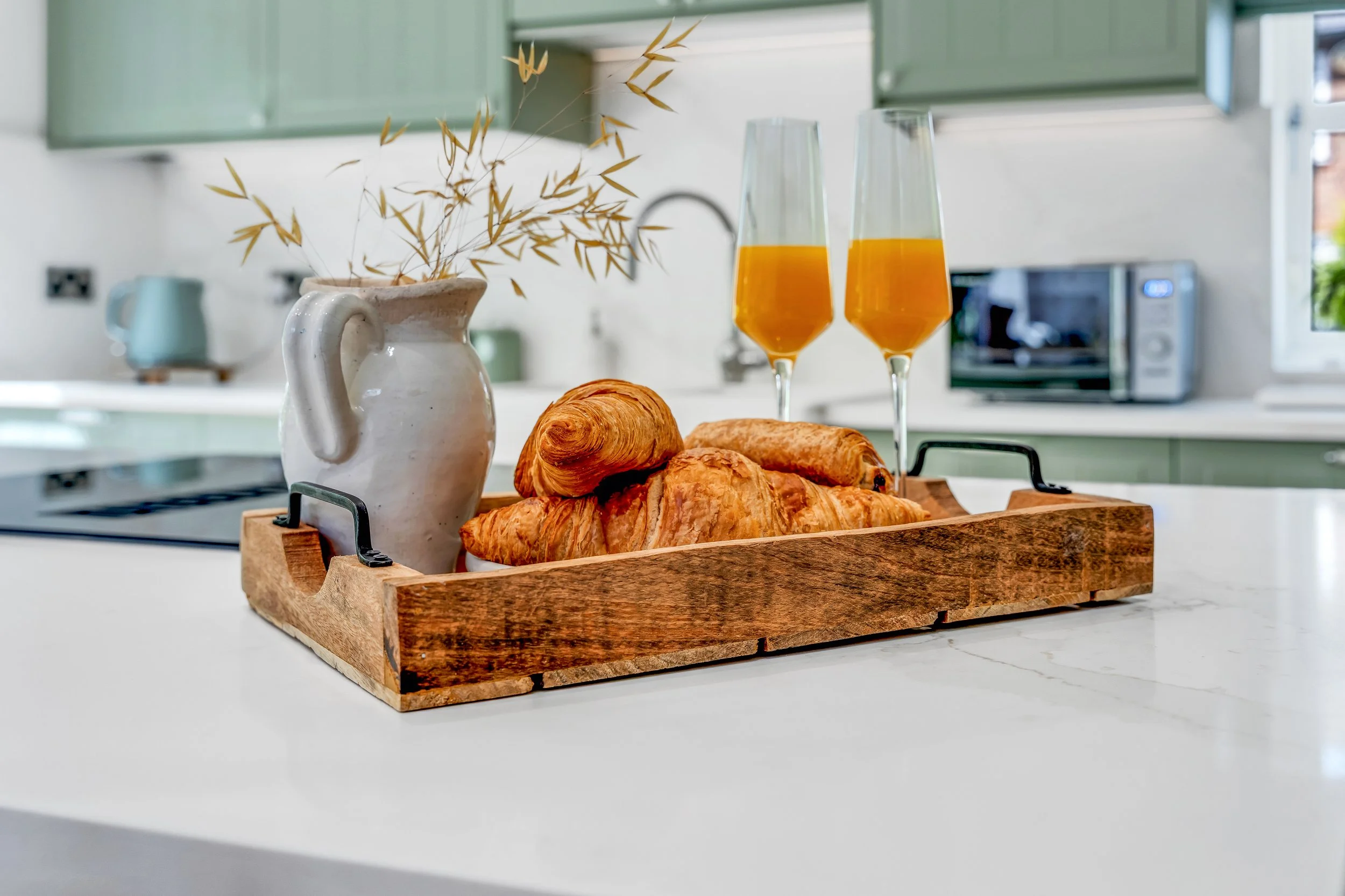 A wooden tray on a white kitchen countertop holding two croissants, a ceramic pitcher with dried plant stems, and two glasses of orange juice. In the background, a microwave oven, a small kettle, and green kitchen cabinets are visible.