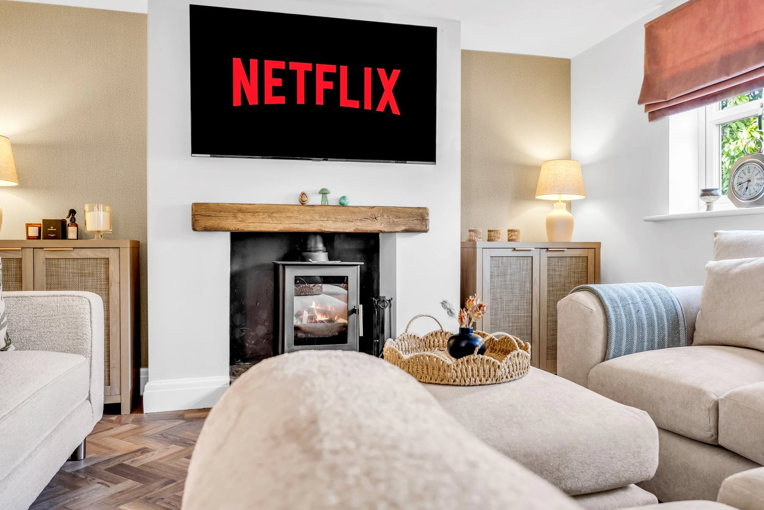 Living room with a wall-mounted TV displaying the Netflix logo, a fireplace with a wooden mantel, beige sofas, side tables with lamps, a window with pink curtains, and decorative items.