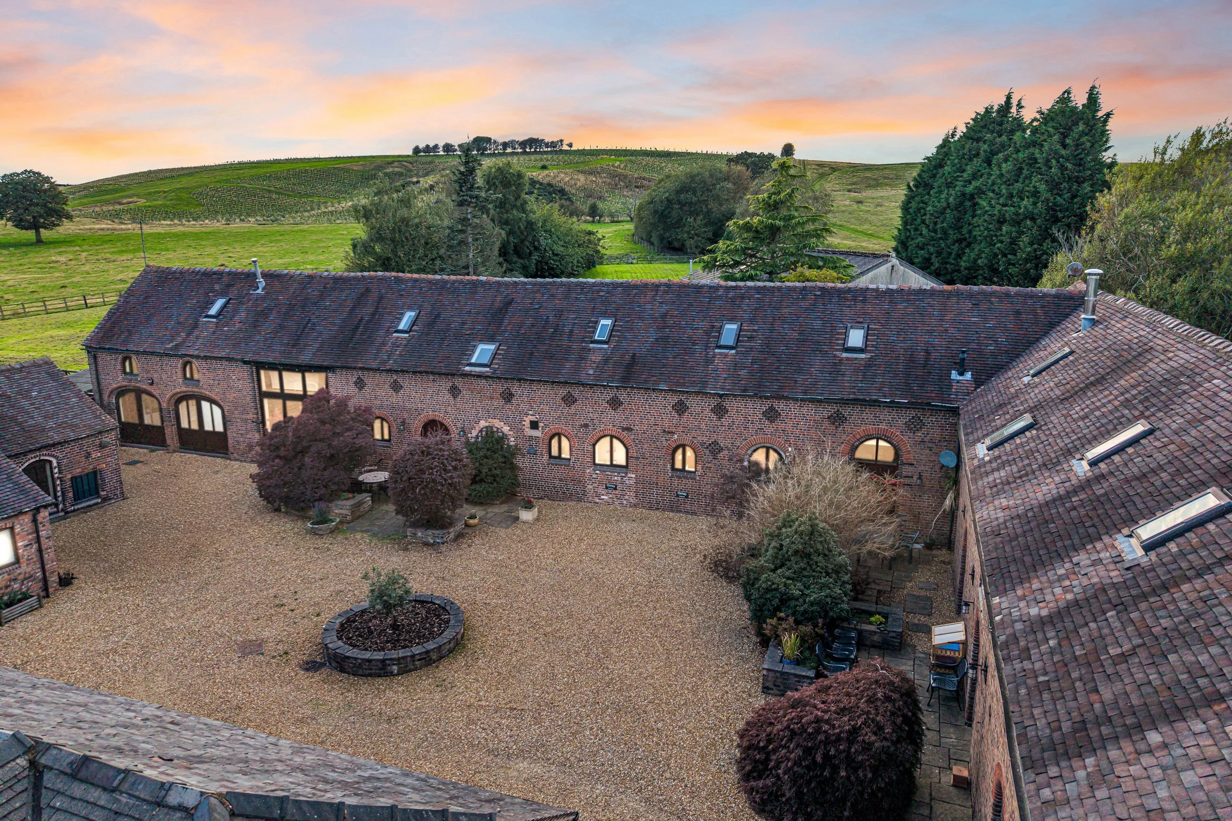A view of a courtyard with brick buildings and a gravel ground, surrounded by trees, with rolling green hills and vineyards in the background during sunset.