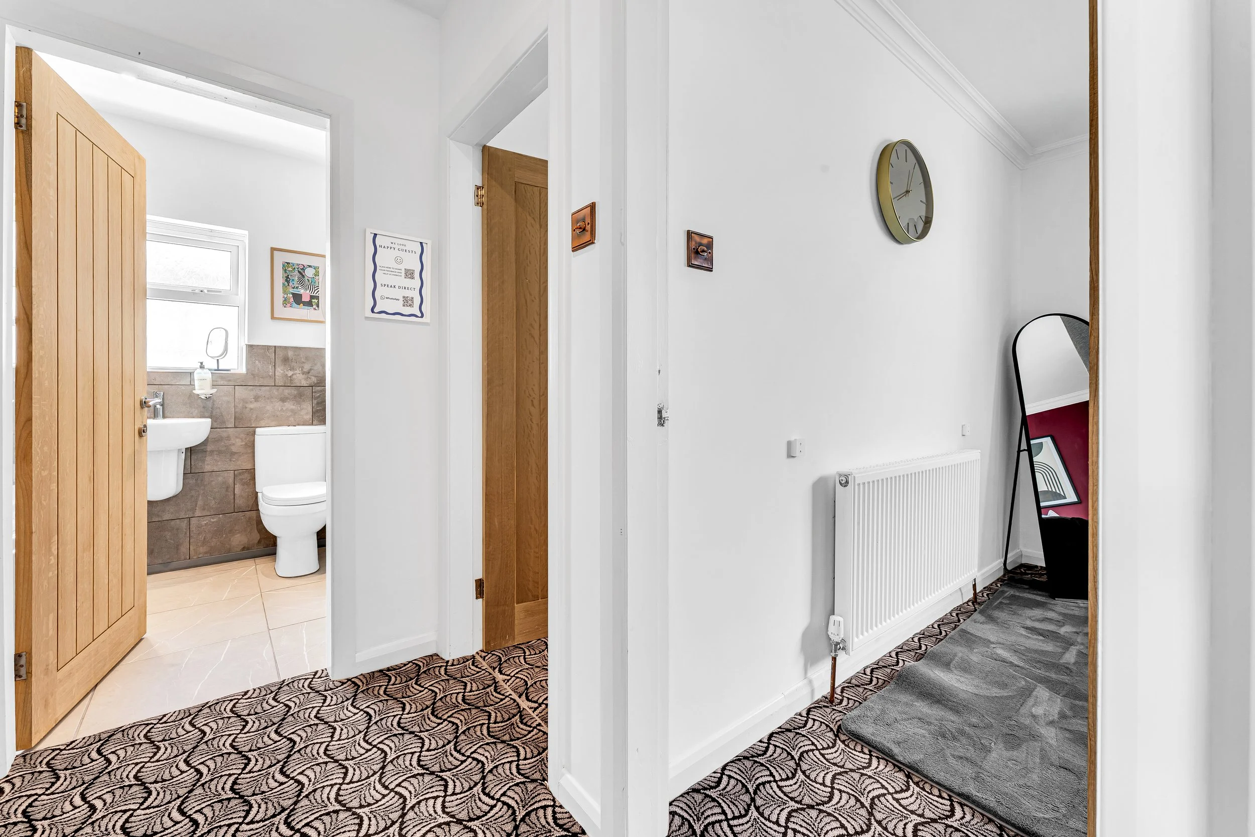 Interior view of a hallway with patterned carpet, leading to a bathroom with wooden door open, showing a toilet and sink, and a mirror and artwork on the wall.