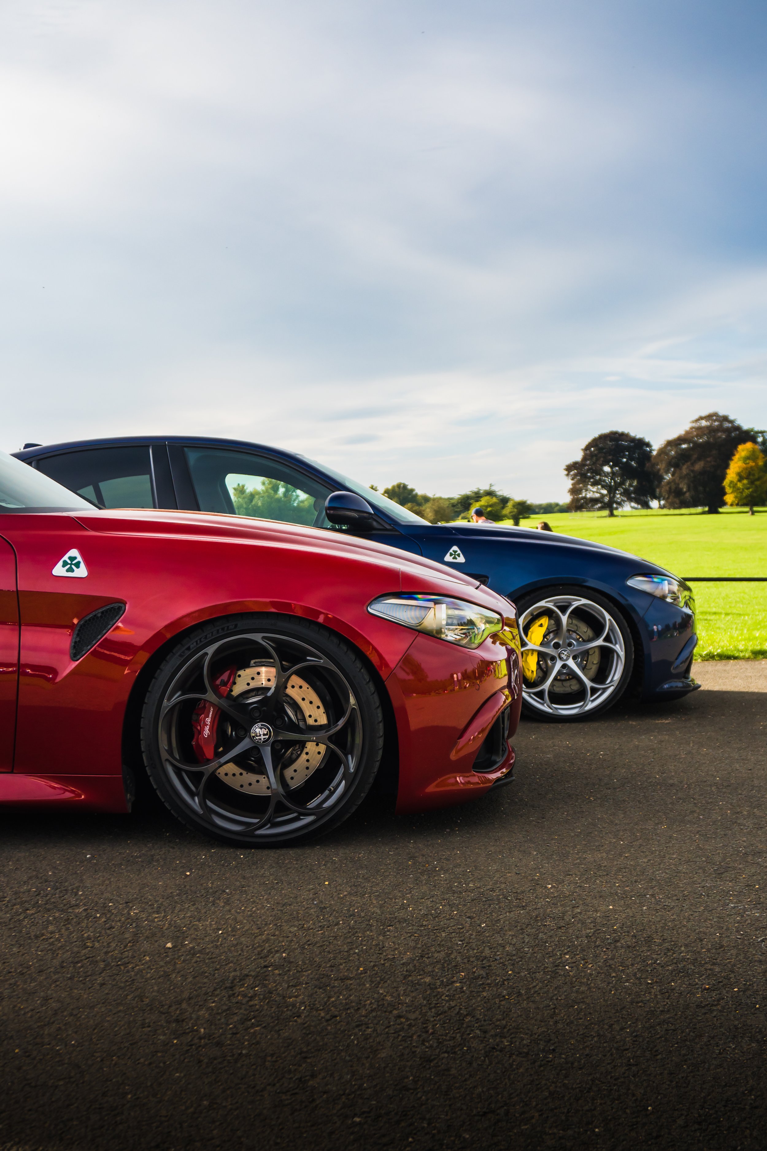 Two sports cars, one red and one dark blue, parked outdoors on a paved surface with a grassy field and trees in the background under a partly cloudy sky.