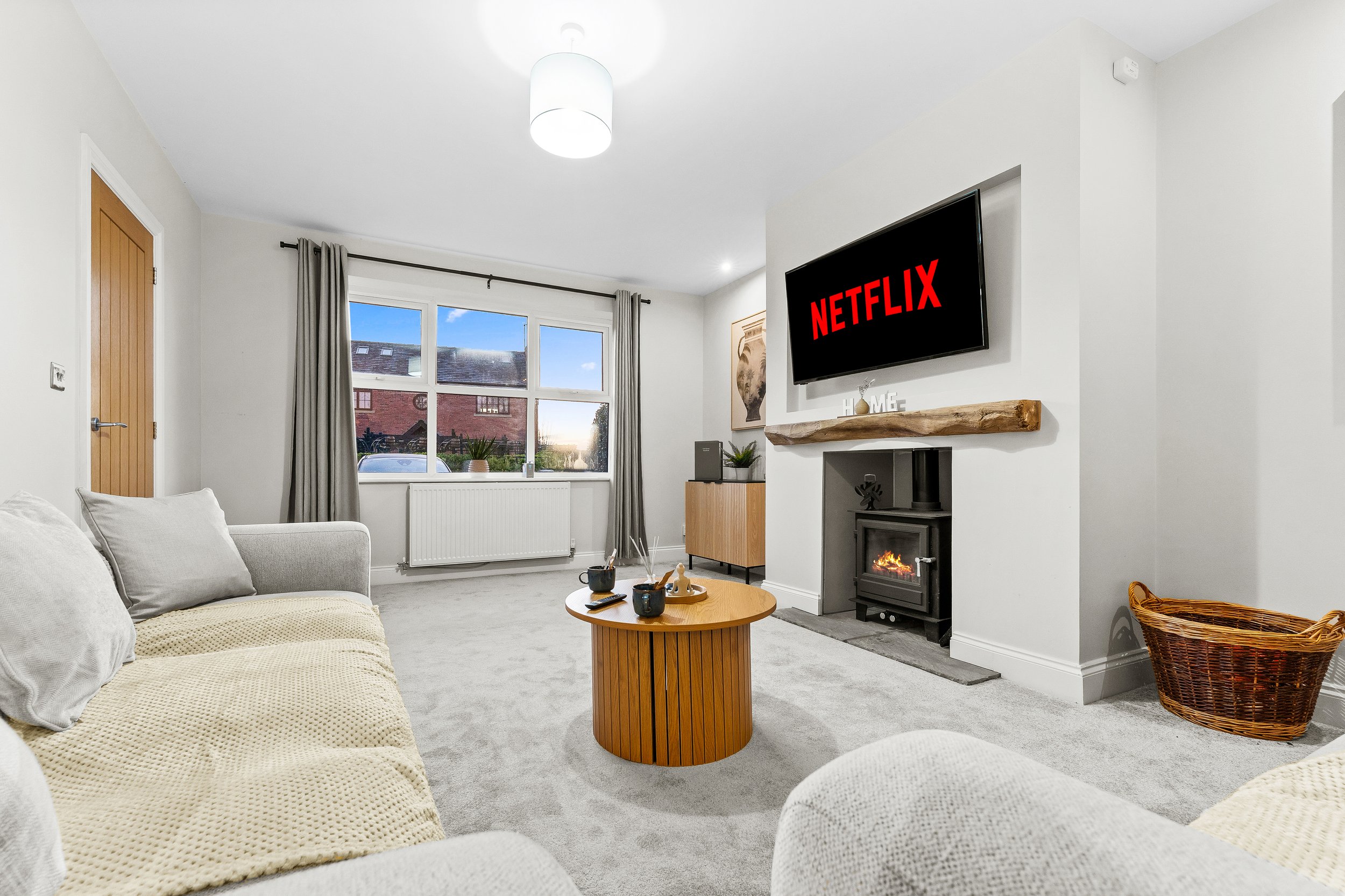 Bright living room with large window, gray curtains, beige couches, wooden coffee table, electric fireplace, wall-mounted TV displaying Netflix, decorative items, and a wicker basket.