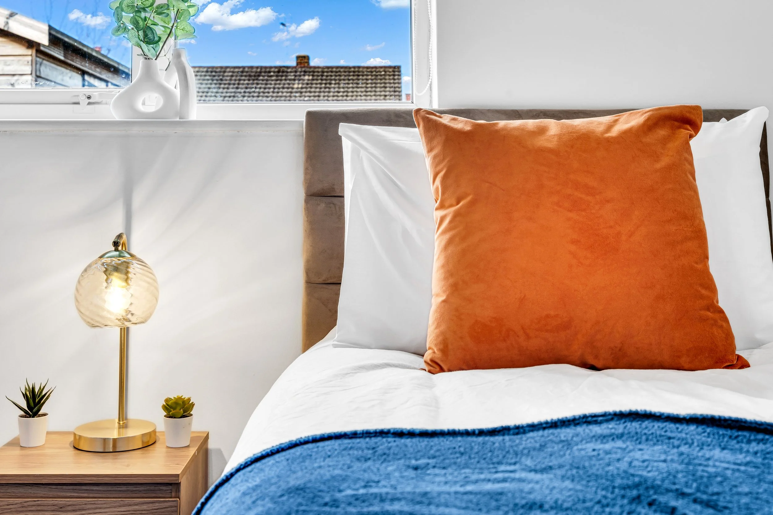 A bedroom with a bed featuring white sheets, an orange pillow, and a blue blanket. A wooden bedside table with a lamp and two small potted plants. A window shows a blue sky and rooftops outside.