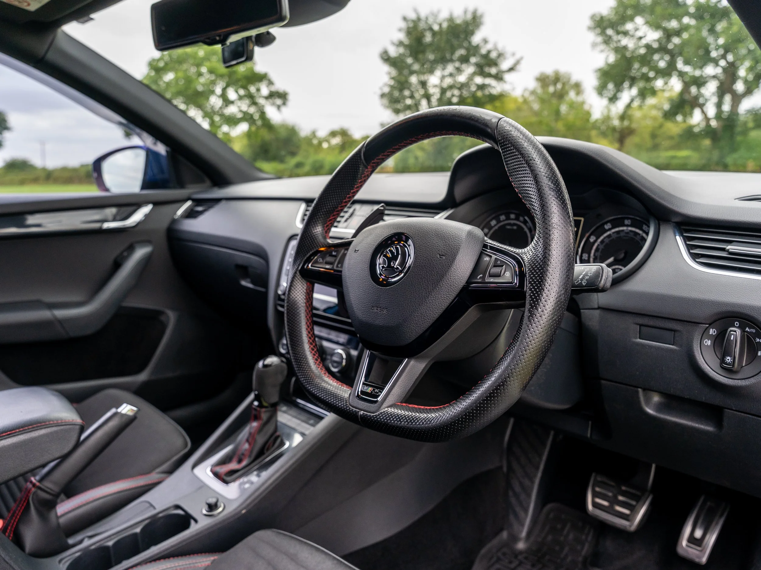 Interior of a car showing the steering wheel, dashboard, gear shift, and side mirror, with a view of trees and sky outside.