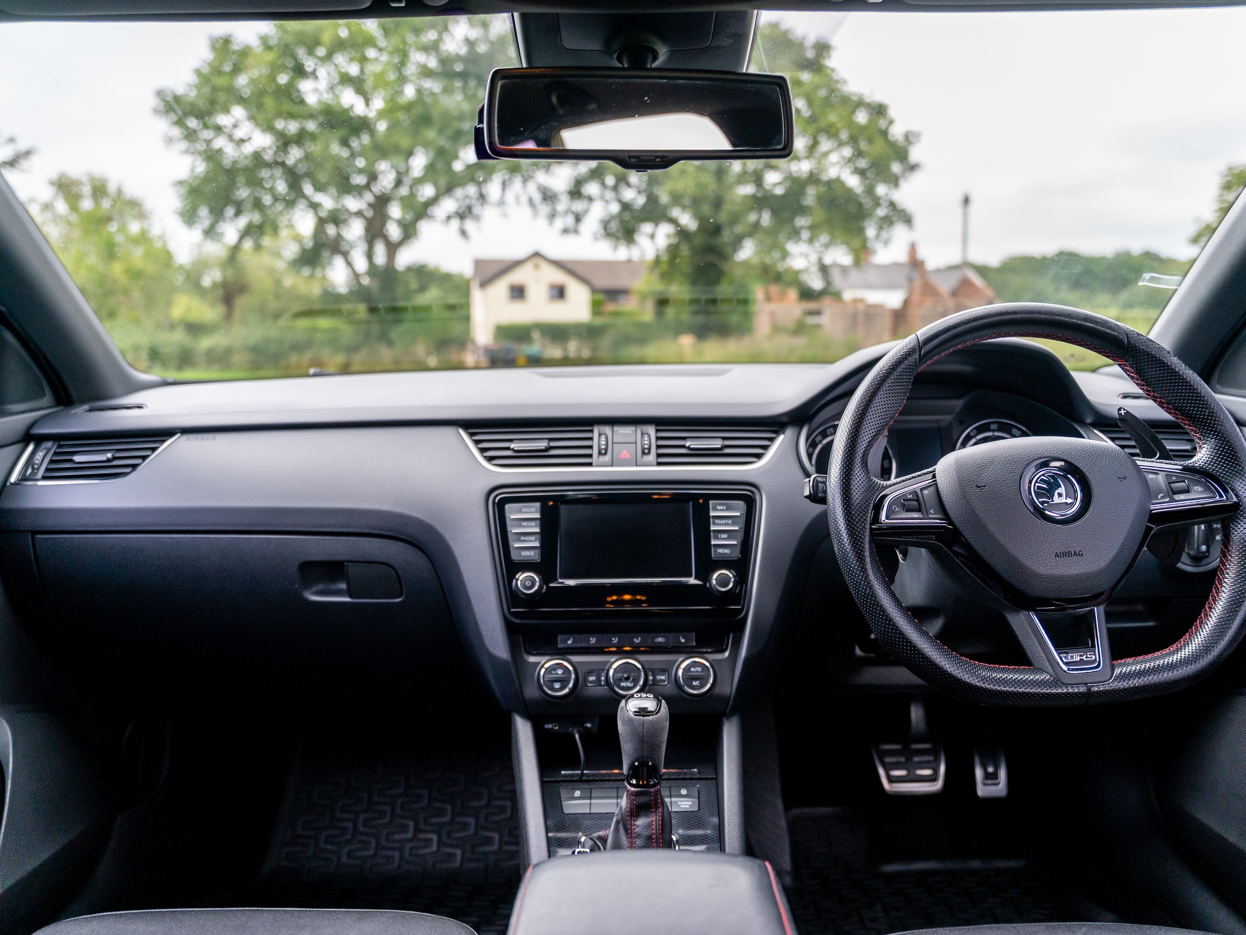 Interior view of a car dashboard with steering wheel, central console, gear shifter, and rearview mirror, with a residential neighborhood visible through the windshield.