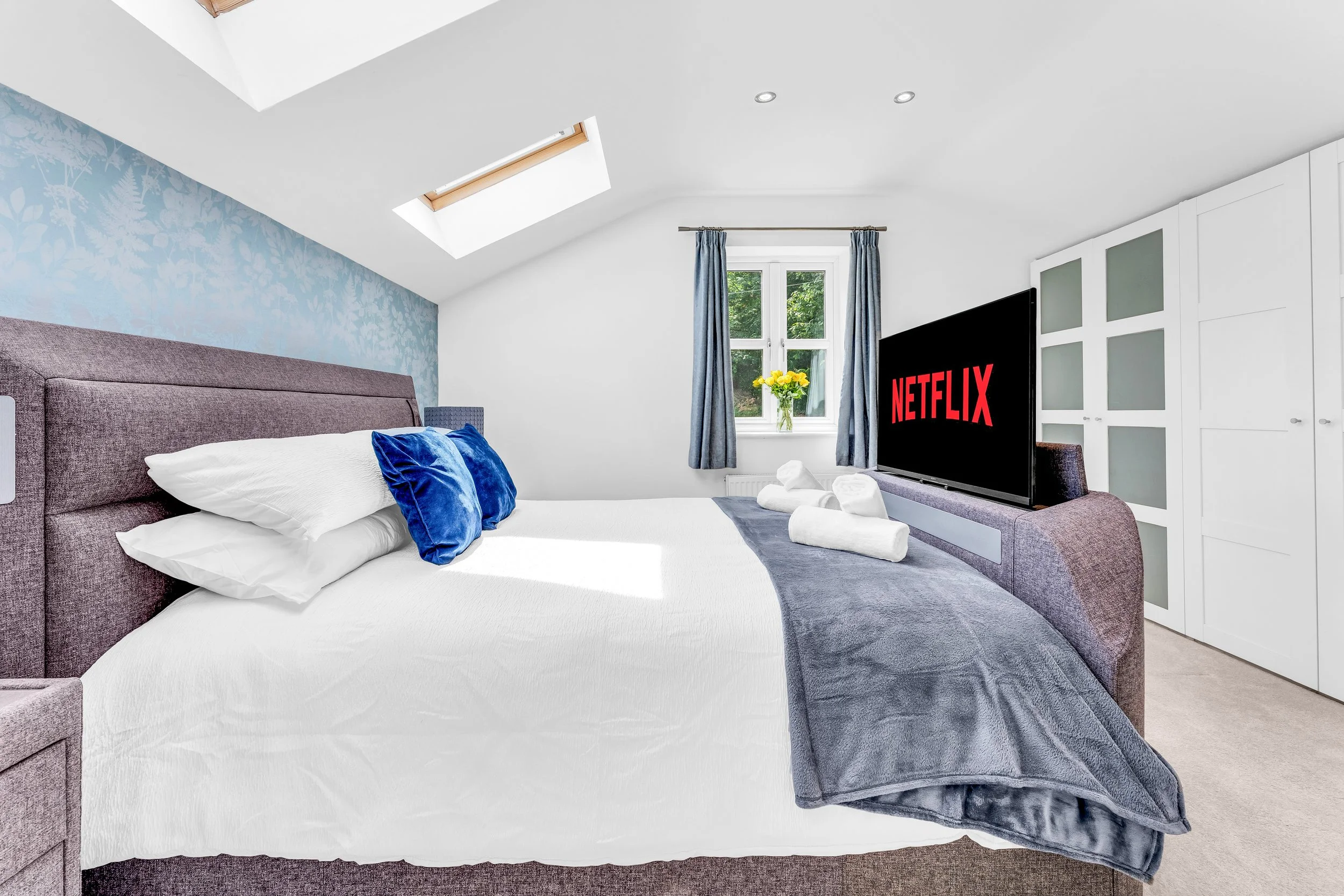 A bedroom with a large bed, white bedding, blue pillows, and a gray blanket. A flat screen TV showing the Netflix logo is on a dresser. There are windows with blue curtains, and a vase with yellow flowers on the windowsill. Skylights in the ceiling p