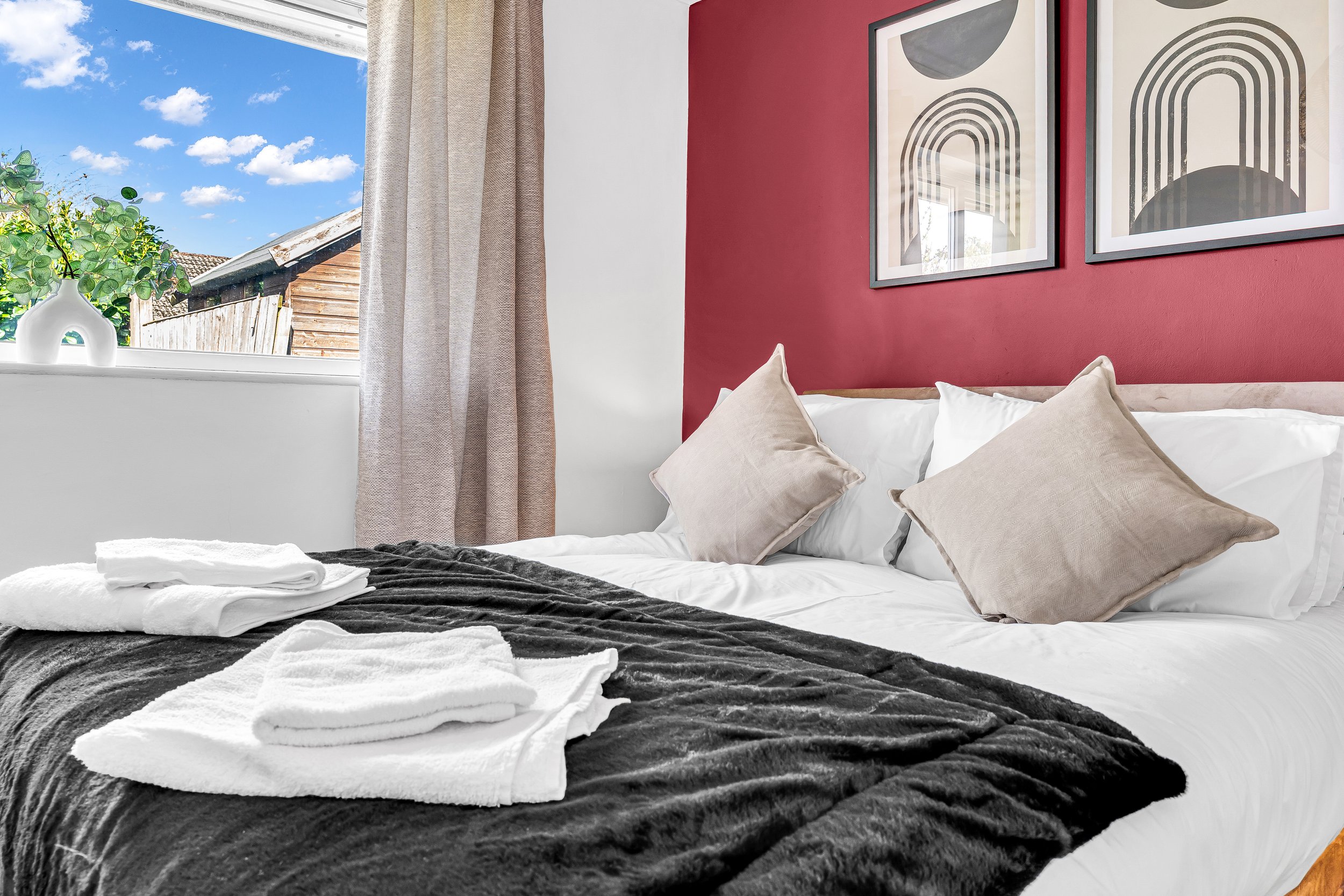 Bedroom with a bed, beige pillows, white sheets, black blanket, and neatly folded towels. Window with beige curtains, outside view of a blue sky and wooden shed, framed abstract art on a red wall.