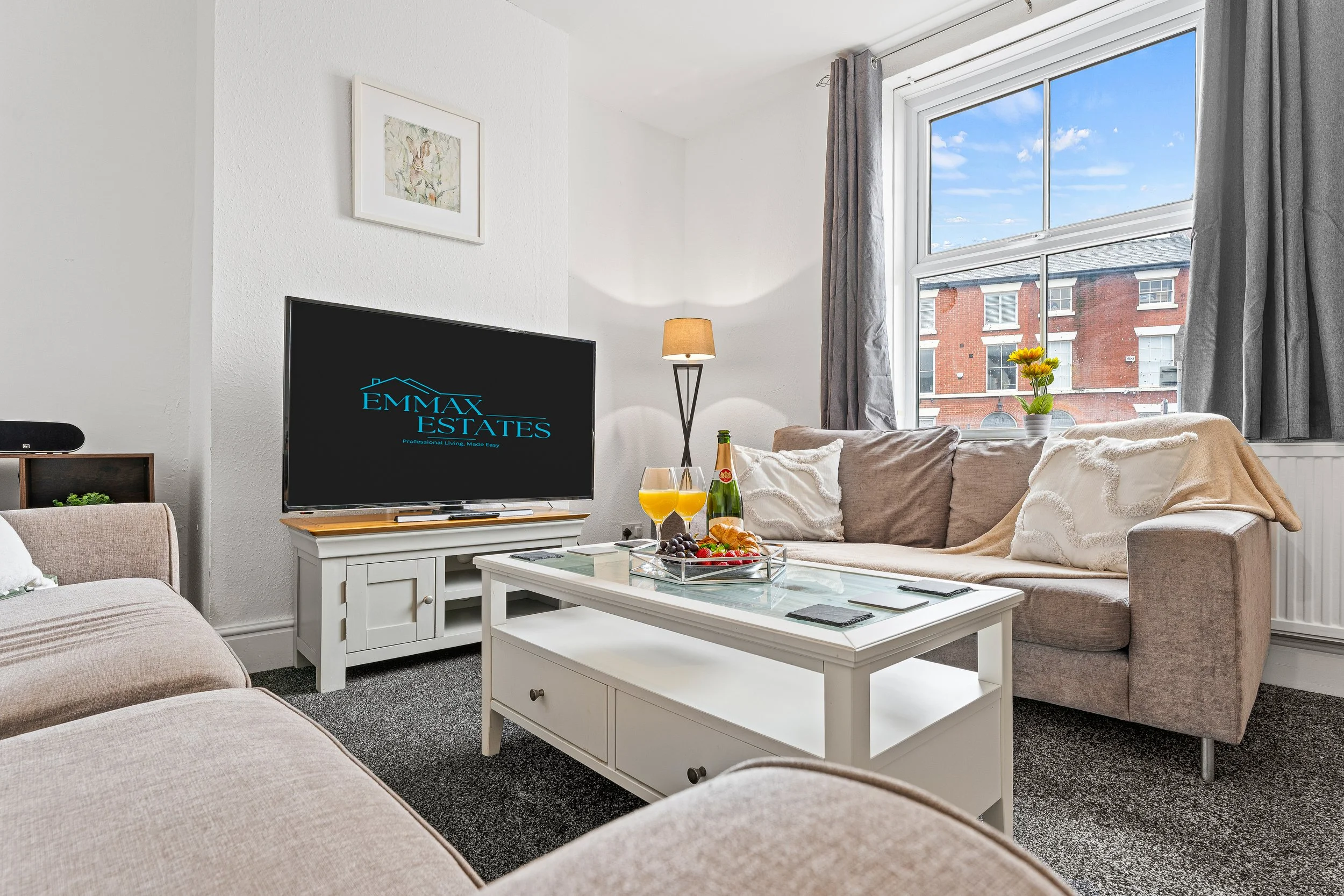 Living room with beige sofas, white coffee table with drinks and snacks, TV on white stand, window with gray curtains showing a brick building and blue sky.