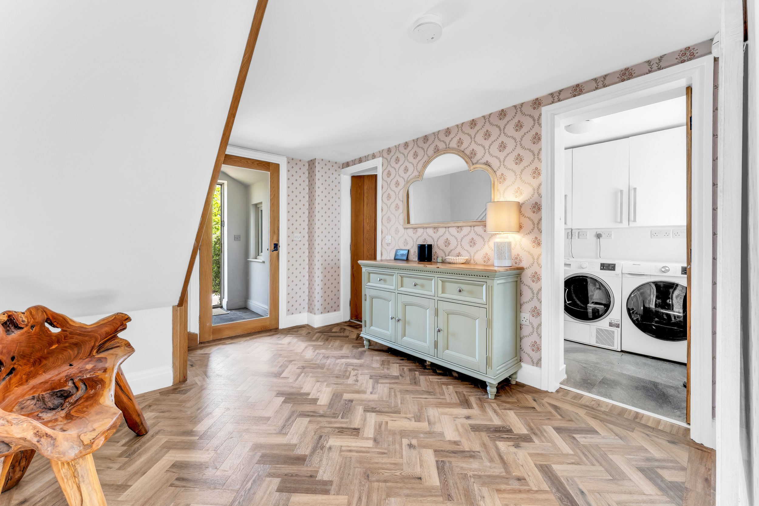 A living space with herringbone hardwood floor, a wooden rock-shaped side table, a mint green sideboard with a mirror, a lamp, and a small electronic device. Visible doorway to a laundry area with washing machines, a door to the outside, and wallpape