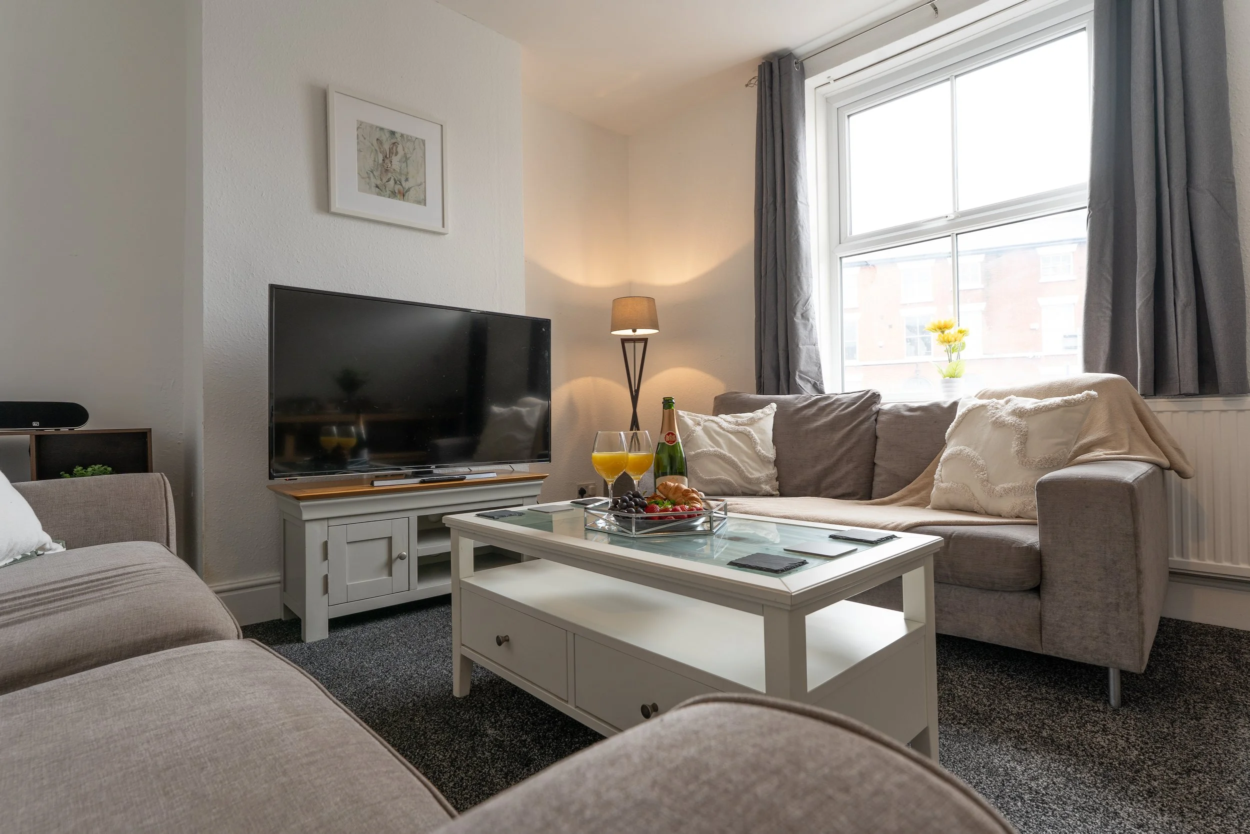 Living room with beige couches, a glass coffee table with snacks and drinks, a television on a stand, a window with curtains, and a floor lamp.