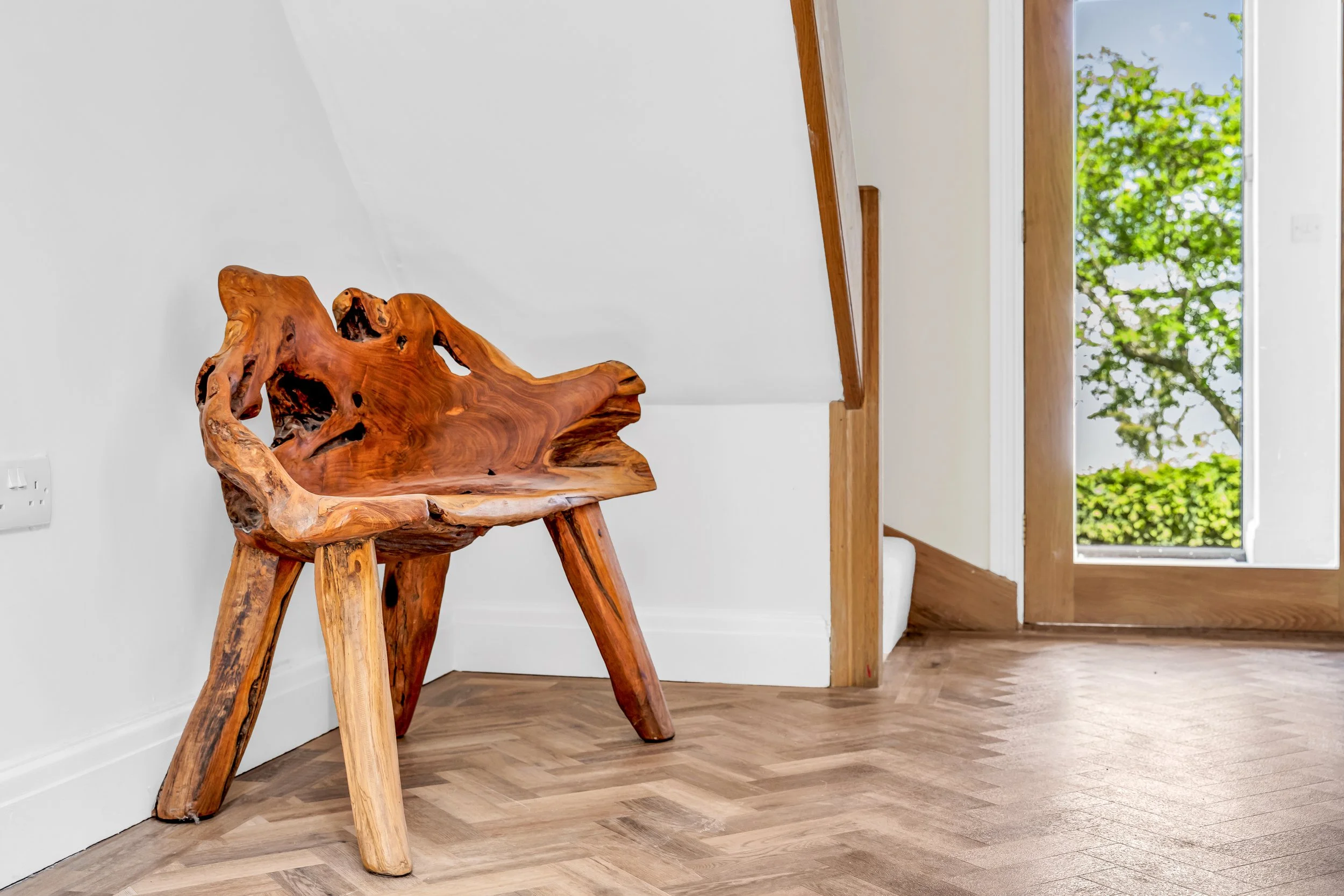 Wooden natural log-style chair in a bright room with a large window showing green trees outside.