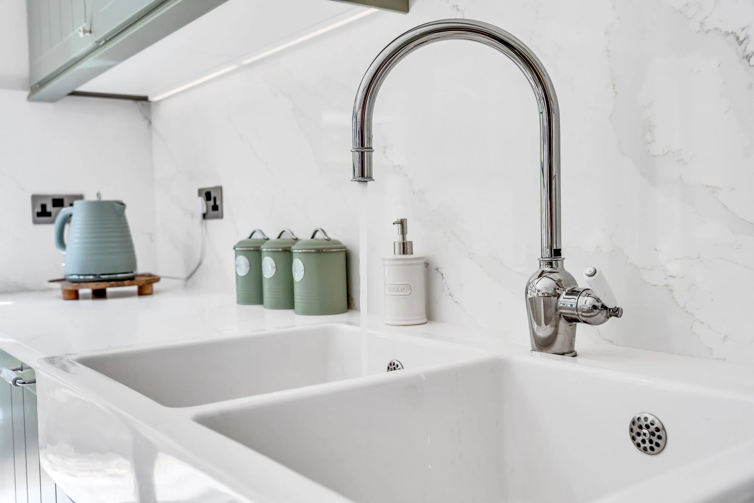 Kitchen sink with a silver faucet, soap dispenser, and green canisters on the counter, with a blue kettle and electrical outlets in the background.