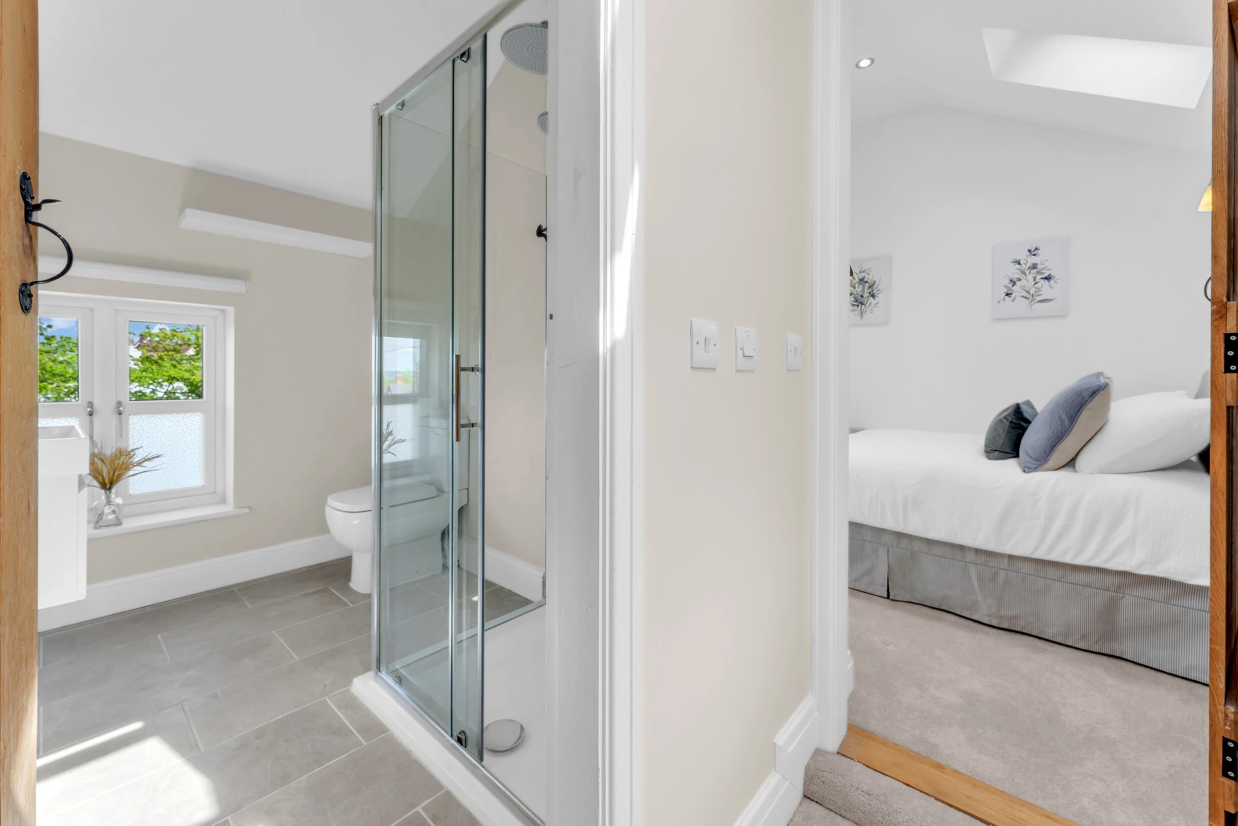 Bathroom with a glass shower stall, toilet, window with view of trees, and a bedroom with a bed and artwork on the wall.