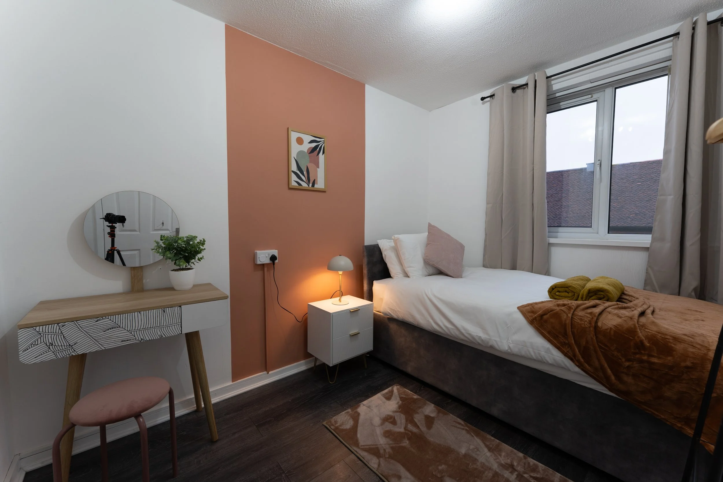 A bedroom with a bed, pillows, folded towels, a desk with a mirror, potted plant, a small nightstand with a lamp, a window with beige curtains, and a pink accent wall.