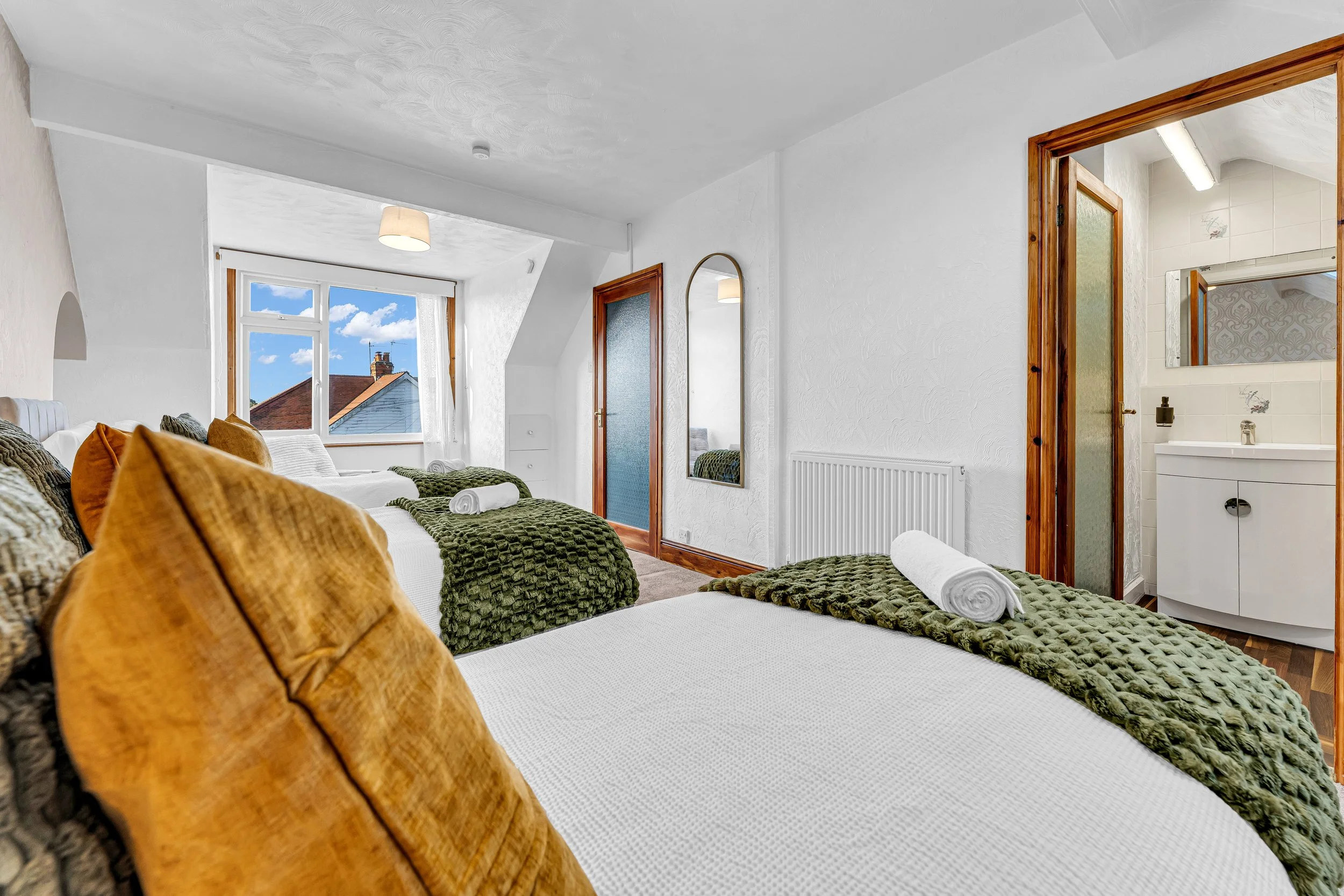 Bedroom with four beds covered in white sheets and green textured blankets, each with rolled towels, and orange throw pillows, large window showing blue sky and rooftops, door leading to small bathroom with sink, mirror, and wallpapered walls, and a mirror on the wall.