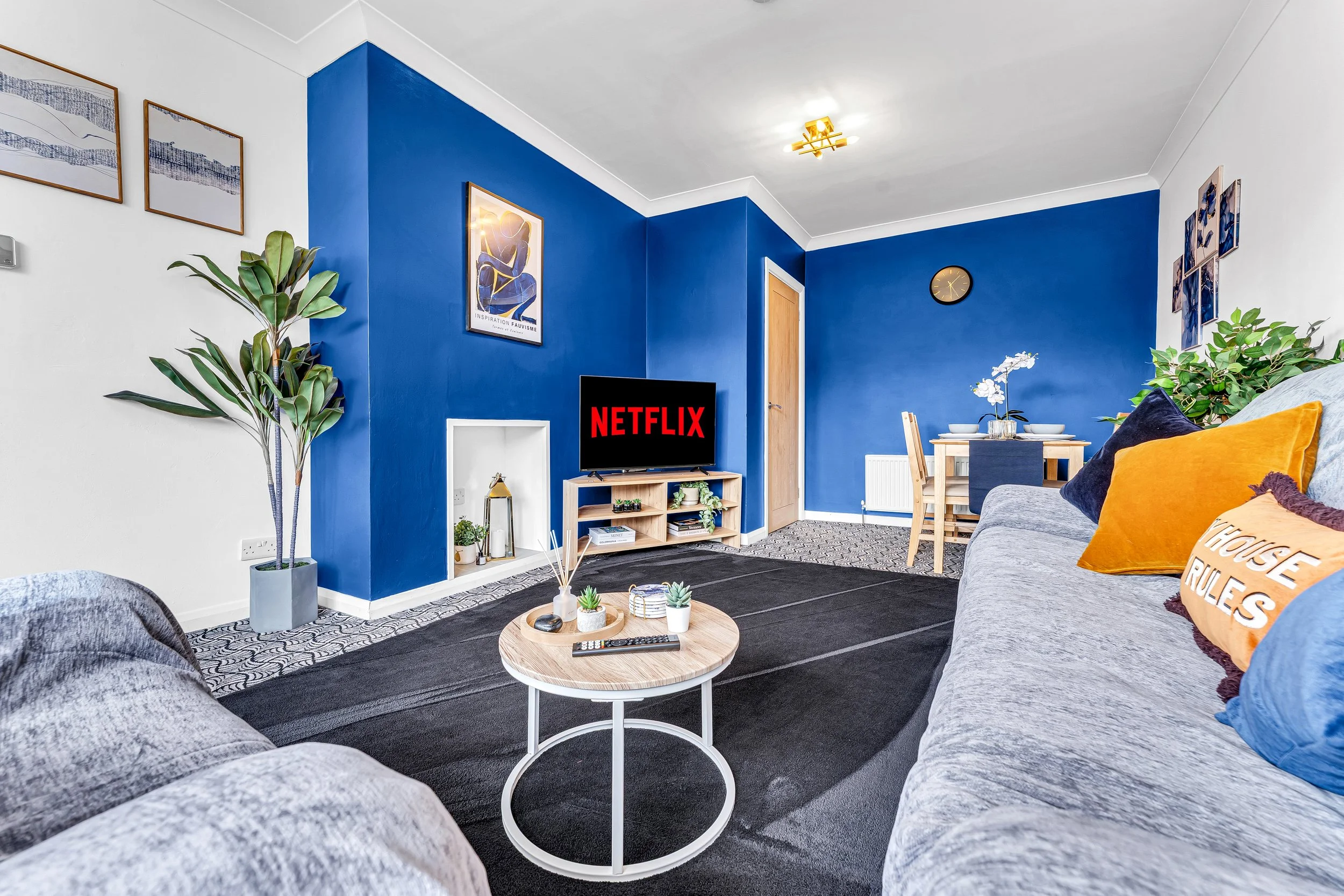 Living room with a gray sofa, a small round coffee table, a TV screen displaying the Netflix logo, a dining table with chairs, a gree plants, and blue accent walls.