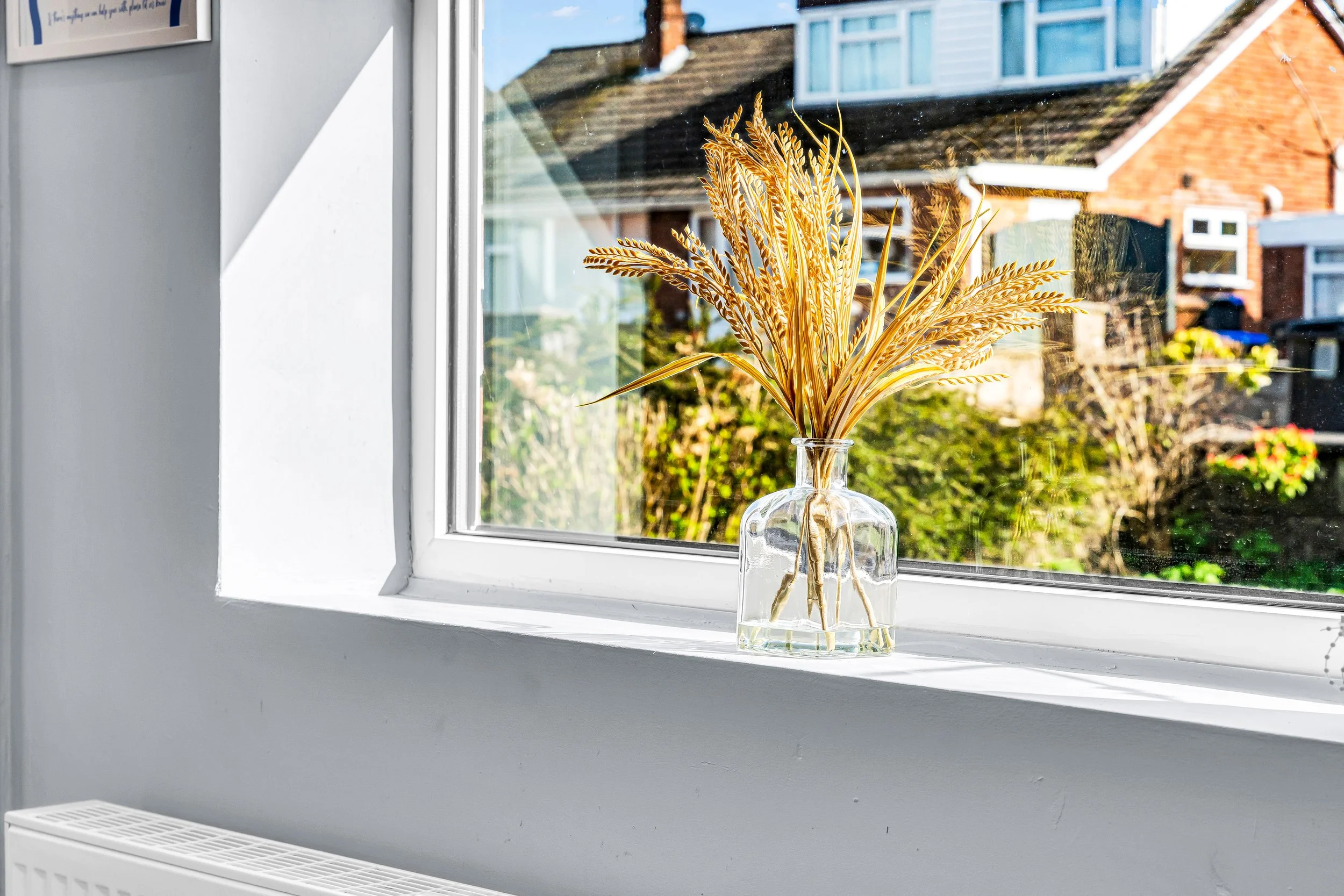 A clear glass vase with dried golden wheat stalks placed on a white window sill in sunlight. Outside view shows houses and greenery.