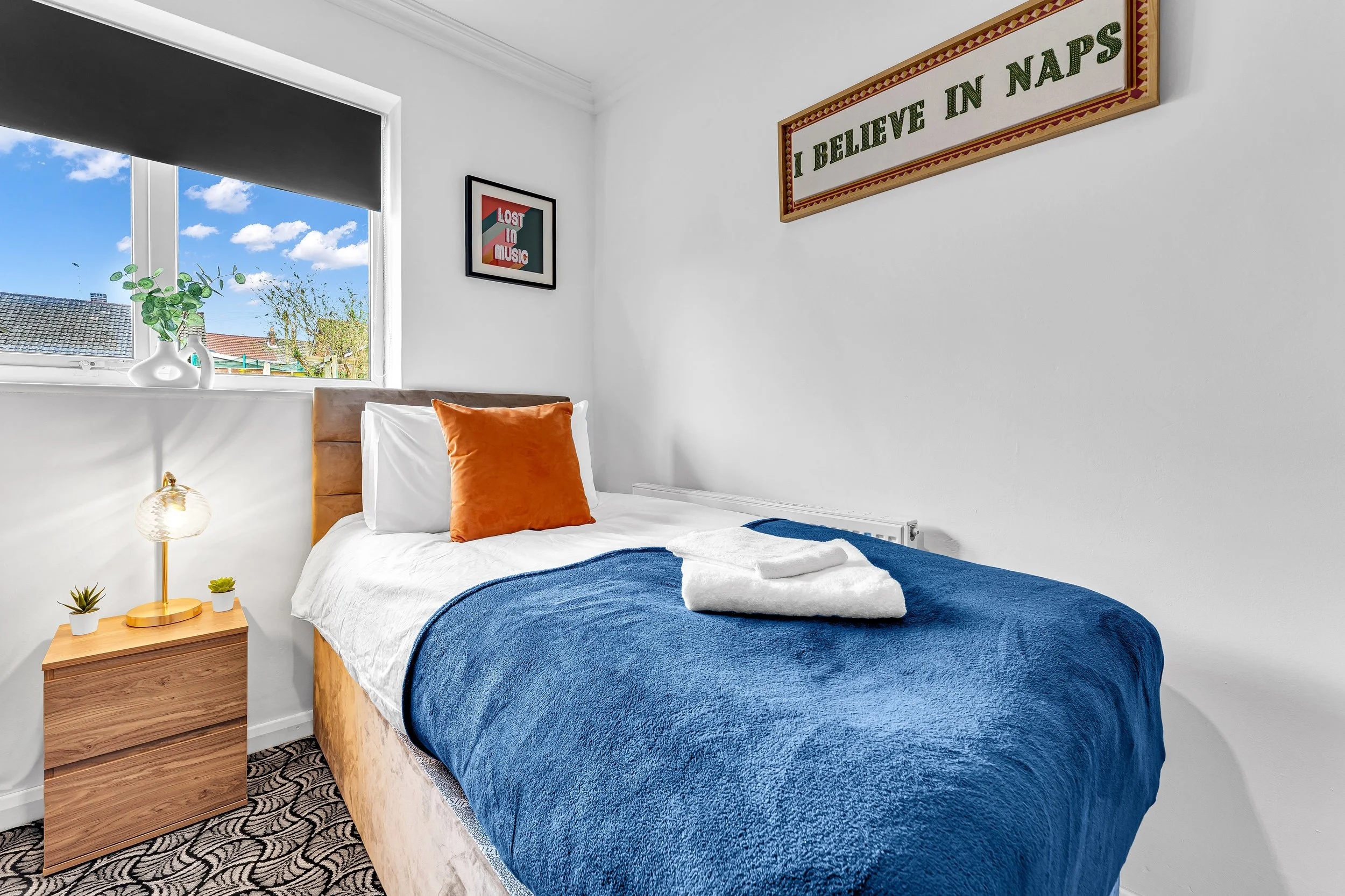 A neatly made bed in a bright bedroom with white walls, a wooden headboard, a blue blanket, white sheets, a white pillow with an orange cushion, a window with a view of rooftops and blue sky, a nightstand with a lamp and small potted plants, and fram