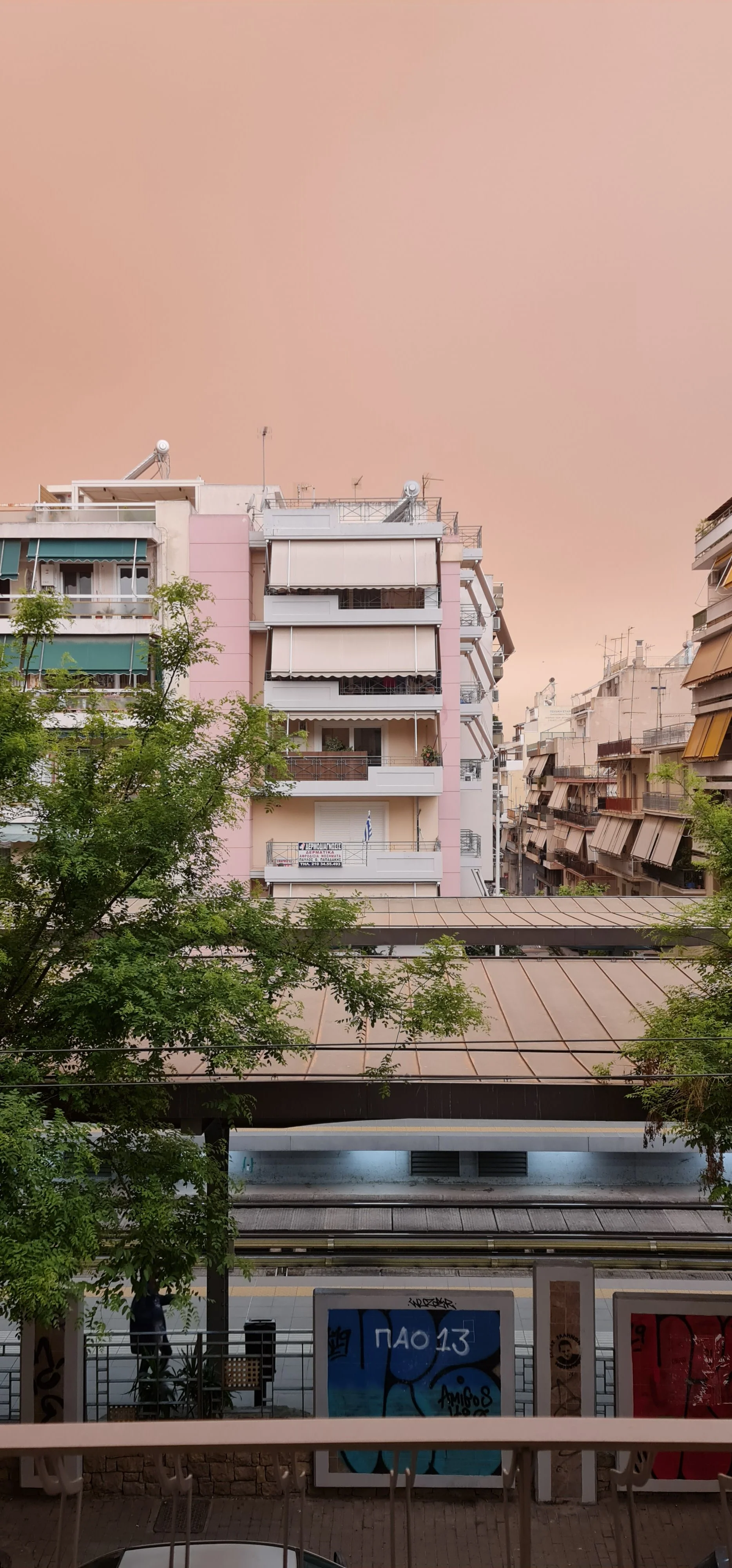 View of a pink apartment building with balconies and awnings, adjacent to other residential buildings, with green trees in the foreground and a train station platform below.