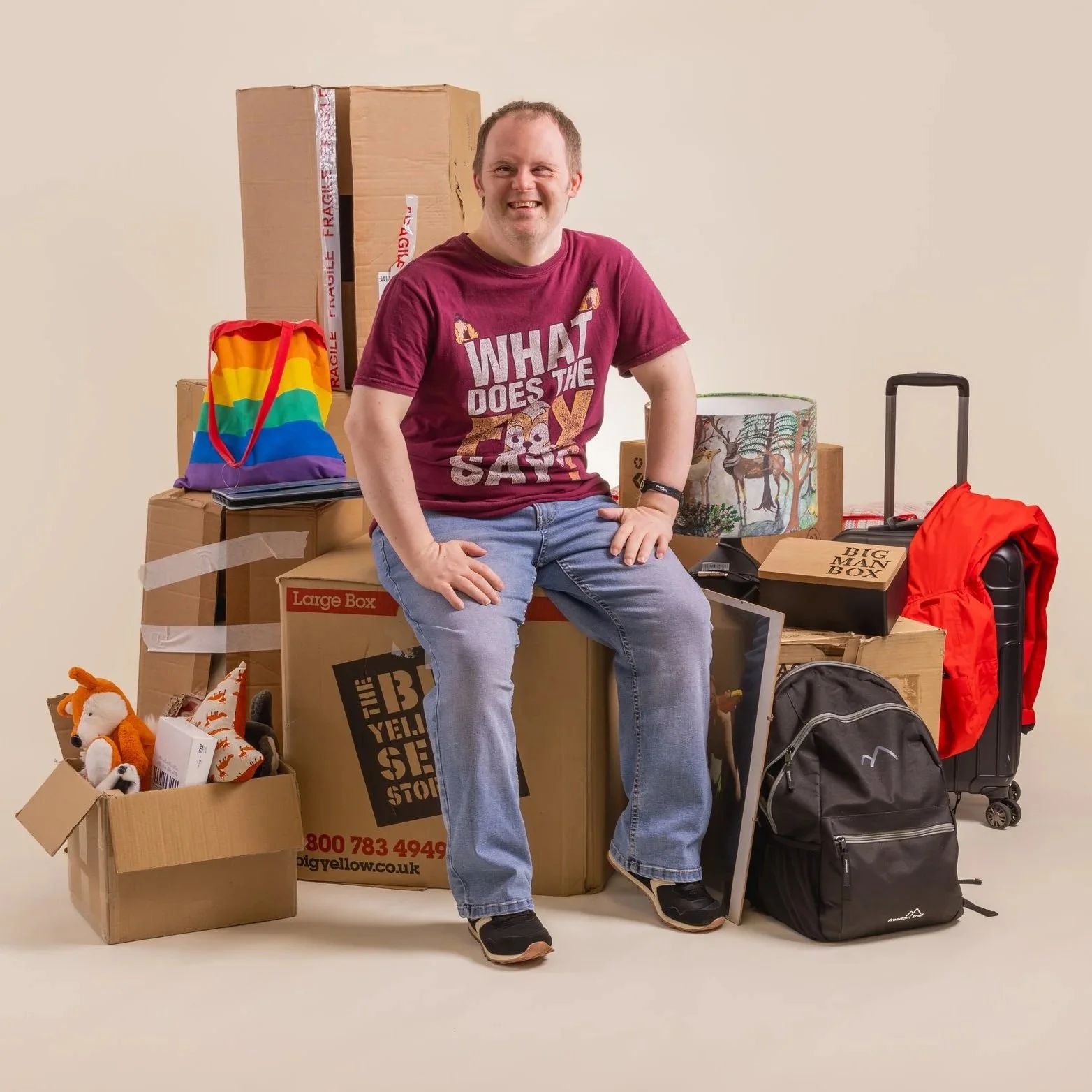 Actor, Nathan Bessell is smiling, sitting on a cardboard box and surrounded by household items and boxes ready to move home. He is wearing a maroon coloured T shirt and blue jeans.